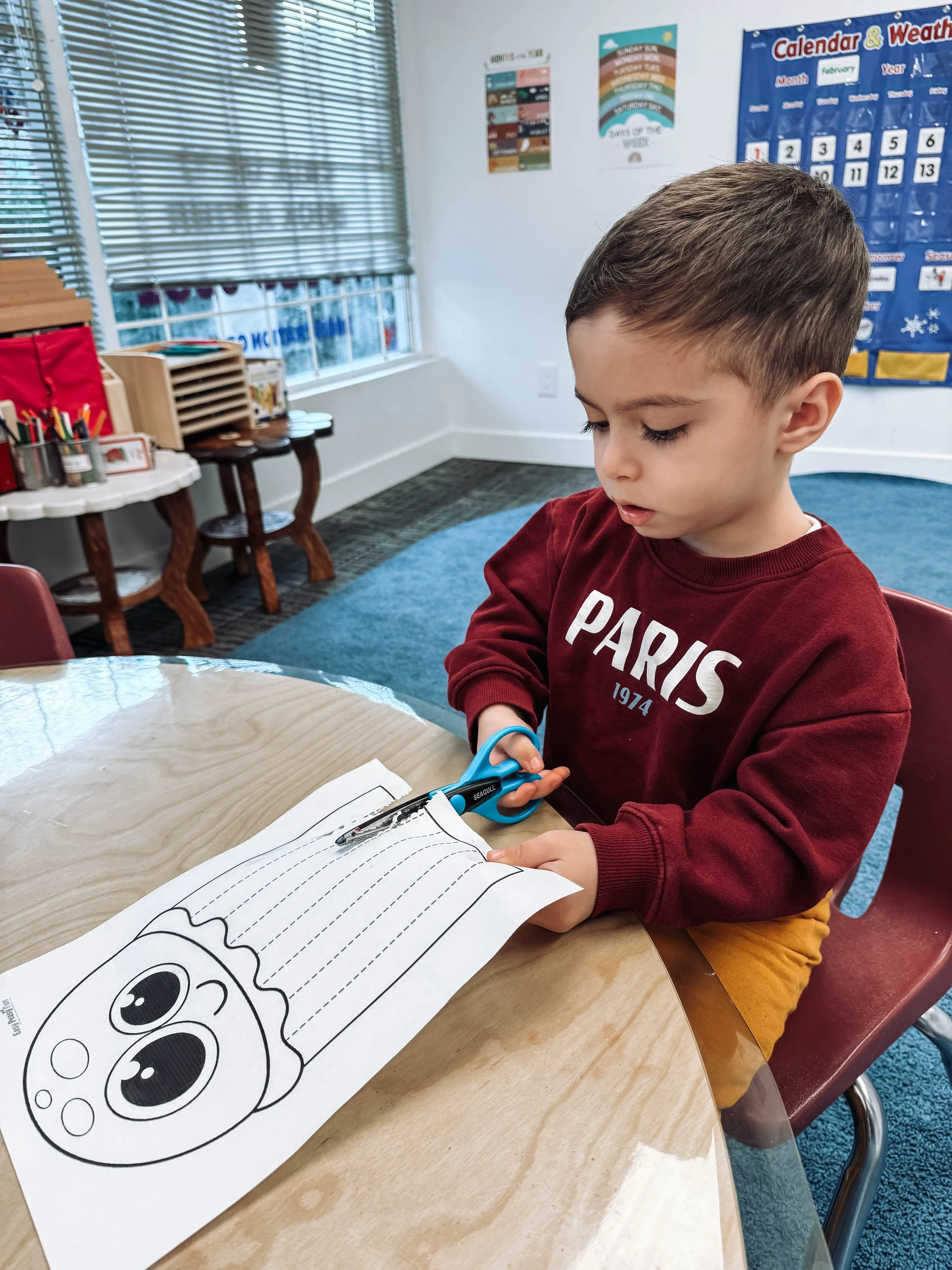 A young boy with brown hair wearing a red sweatshirt with the word 'PARIS' on it, sitting at a wooden table in a classroom, cutting a coloring sheet with scissors. The coloring sheet has a cartoon face of a cupcake or similar dessert. The classroom h