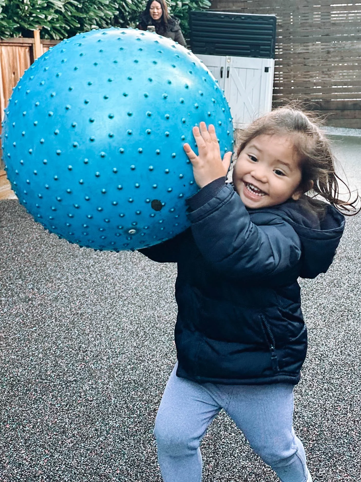 A young girl with curly hair smiling and wearing a black jacket and grey pants, holding a large blue spiky ball, outside on a textured ground with a wooden fence and a woman in the background.