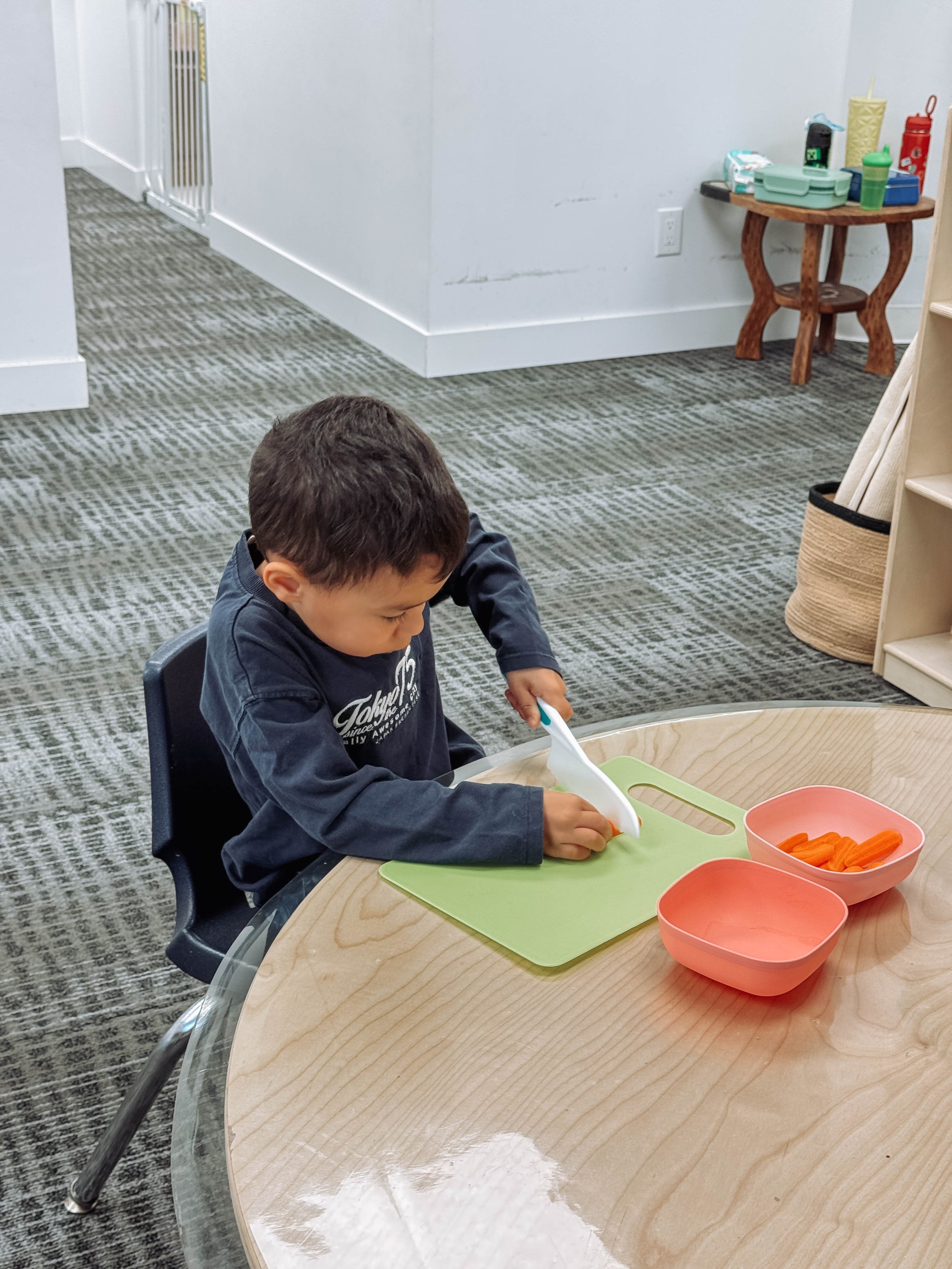 A young boy with dark hair eating baby carrots with a vegetable peeler at a round wooden table.