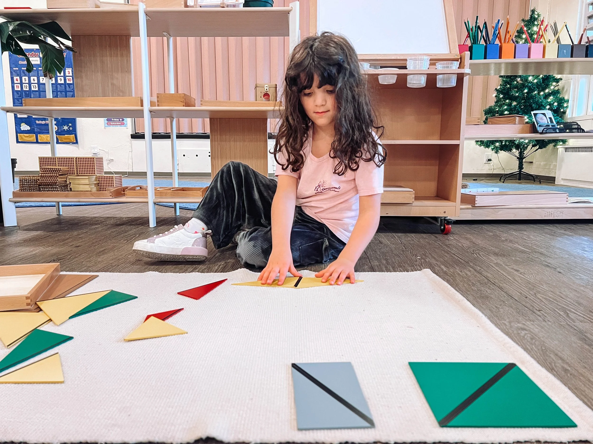 Girl with curly hair sitting on a white carpet, playing with geometric puzzle pieces, in a classroom with shelves and educational materials.