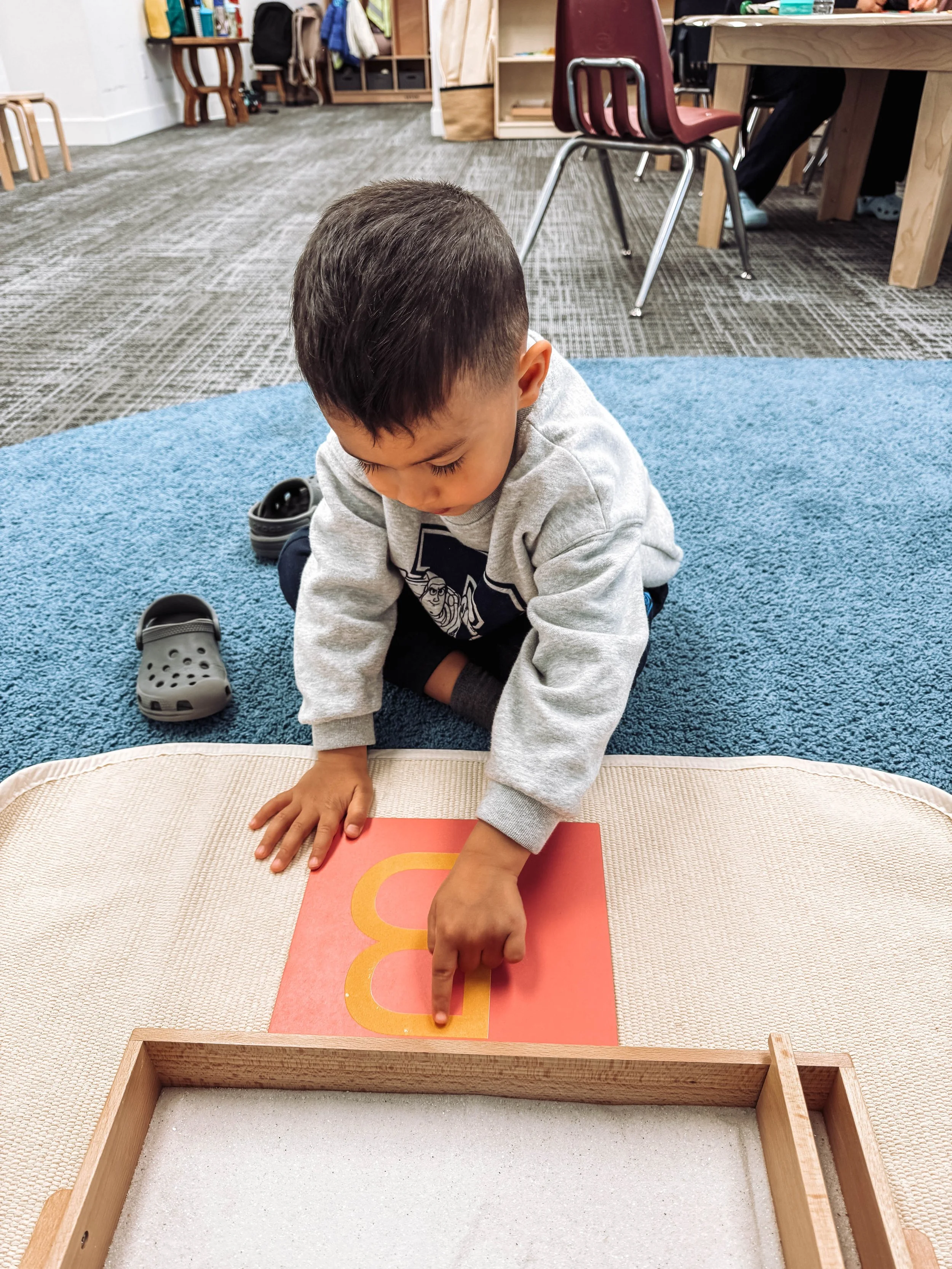 A young boy in a gray sweatshirt and dark pants kneeling on a blue carpet, pointing at a lowercase letter 'b' on a red card.