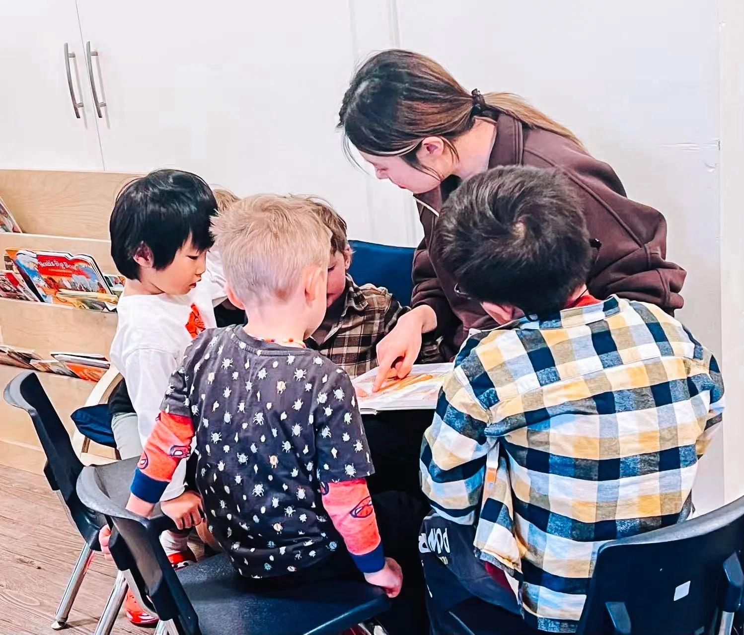 A group of children gathered around a woman, who is showing them something in a book. The children are sitting on chairs, and books are visible on a nearby shelf.