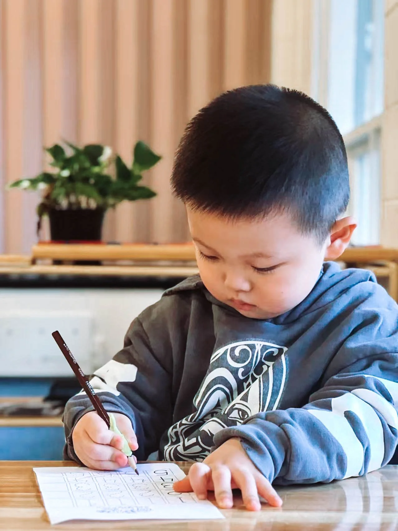 A young boy with short black hair is sitting at a wooden table, writing on a piece of graph paper with a black pen. In the background, there is a potted plant on a wooden shelf, against a backdrop of beige curtains and a window.