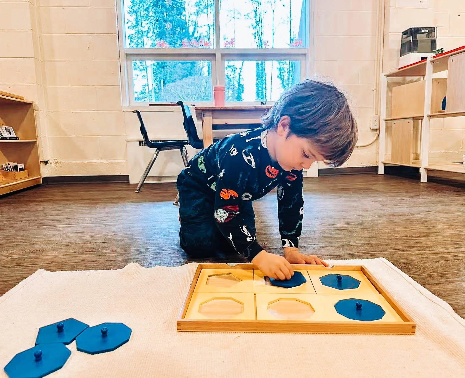 A young boy in a dark printed pajamas is kneeling on a white carpet, playing with a wooden peg game on the floor with blue playing pieces. The room has large windows, wooden furniture, and a wooden floor.
