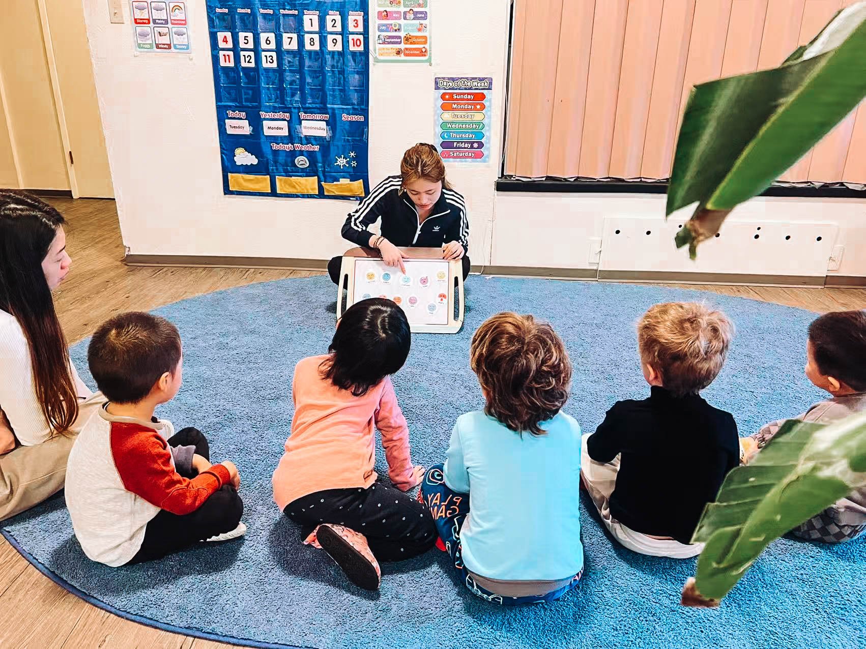 A woman reading a book to a group of five children sitting on a blue rug in a classroom.