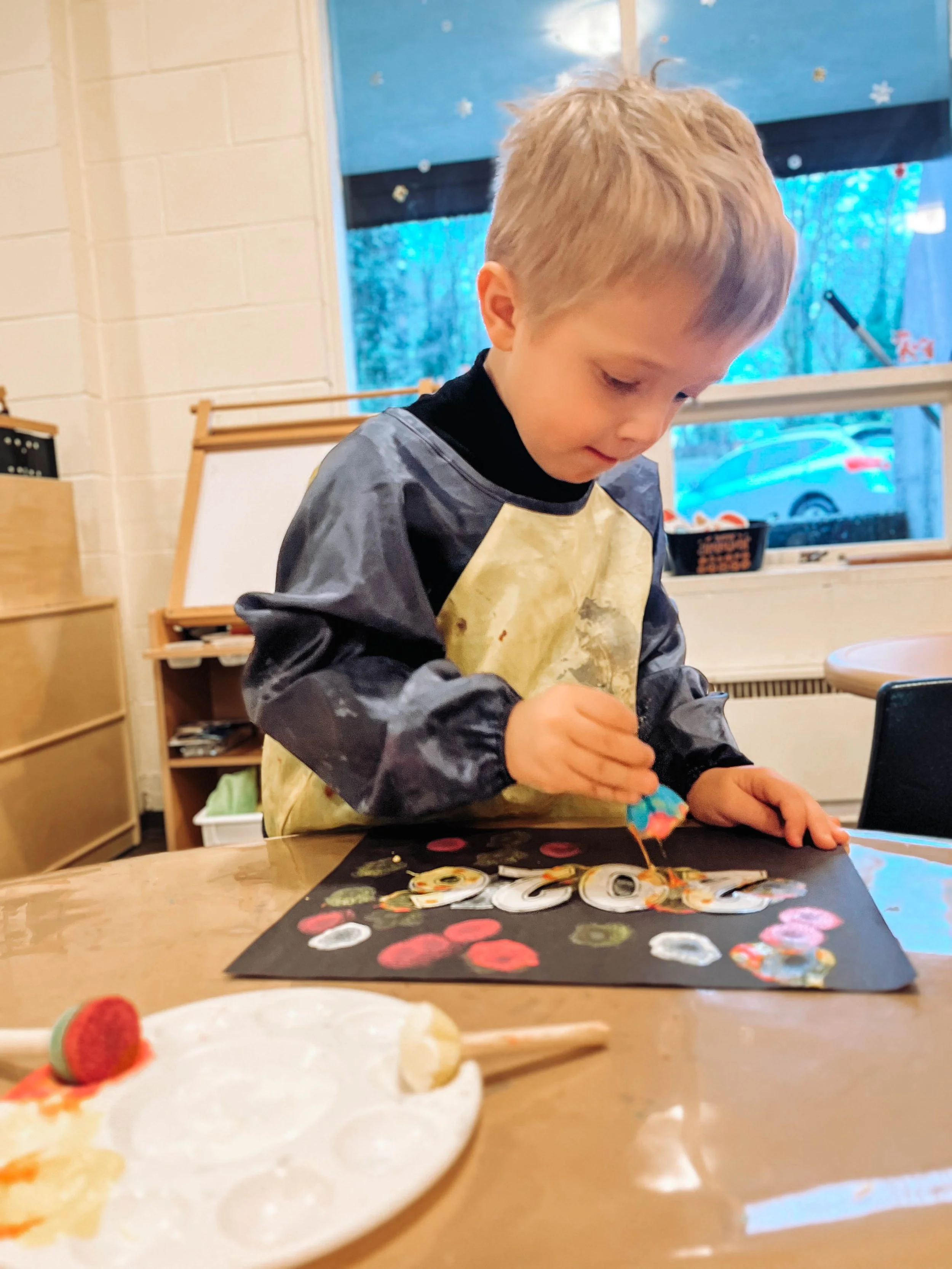 A young boy wearing an art smock is working on an art project at a table. He is glueing or placing embellishments on a black paper with colorful decorations. There is a paint palette with some paint and an eraser in the foreground, and in the background, a window showing trees and a parked car outside.