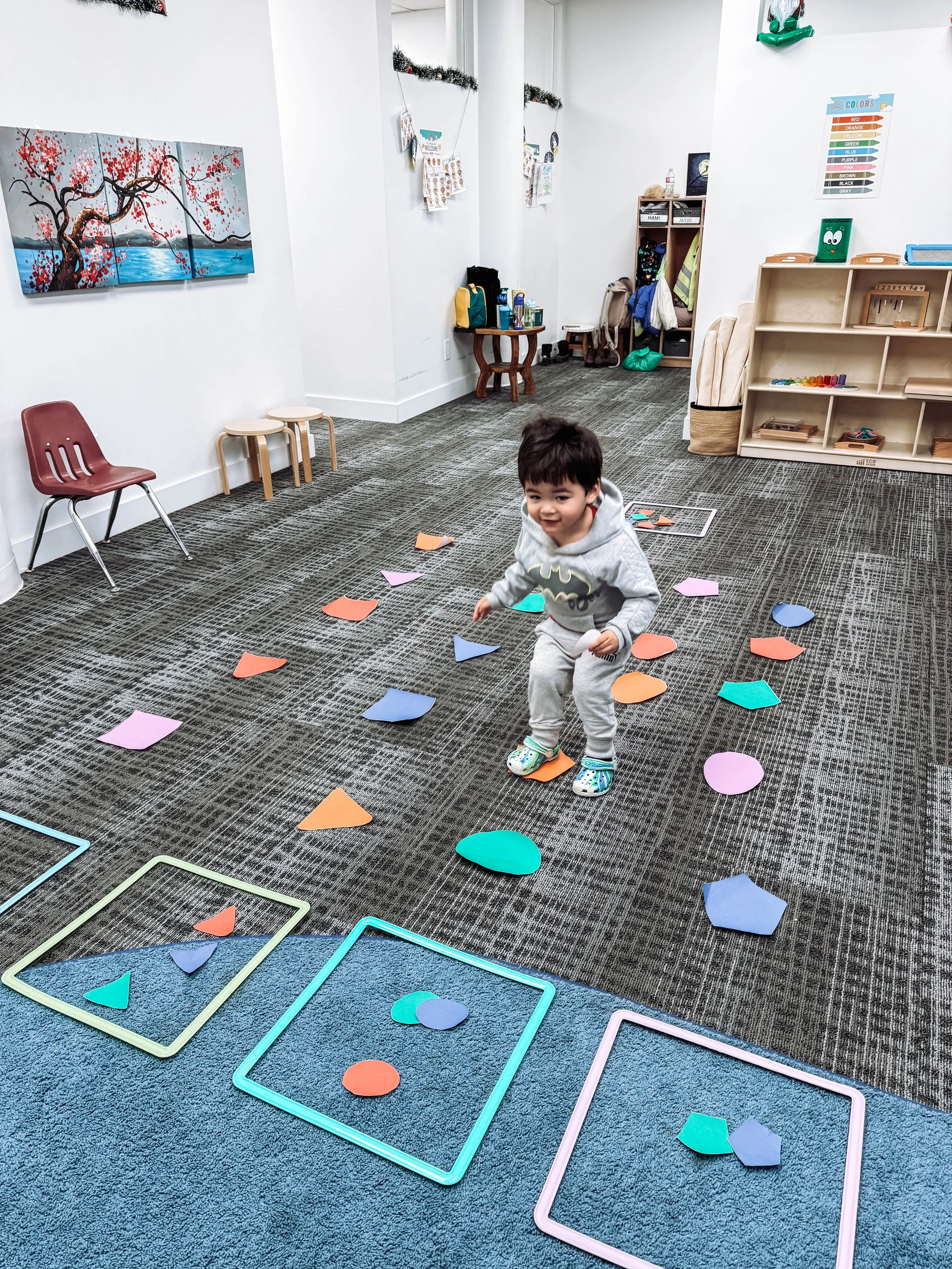 Young boy smiling and playing with colorful paper shapes on the floor of a classroom or playroom, with educational posters and shelves in the background.