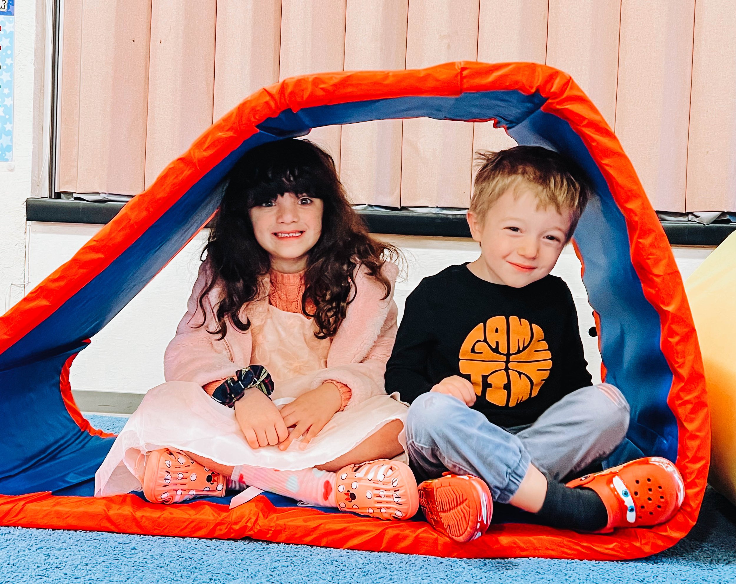 Two children sitting inside a play tunnel on a blue carpet, smiling at the camera.