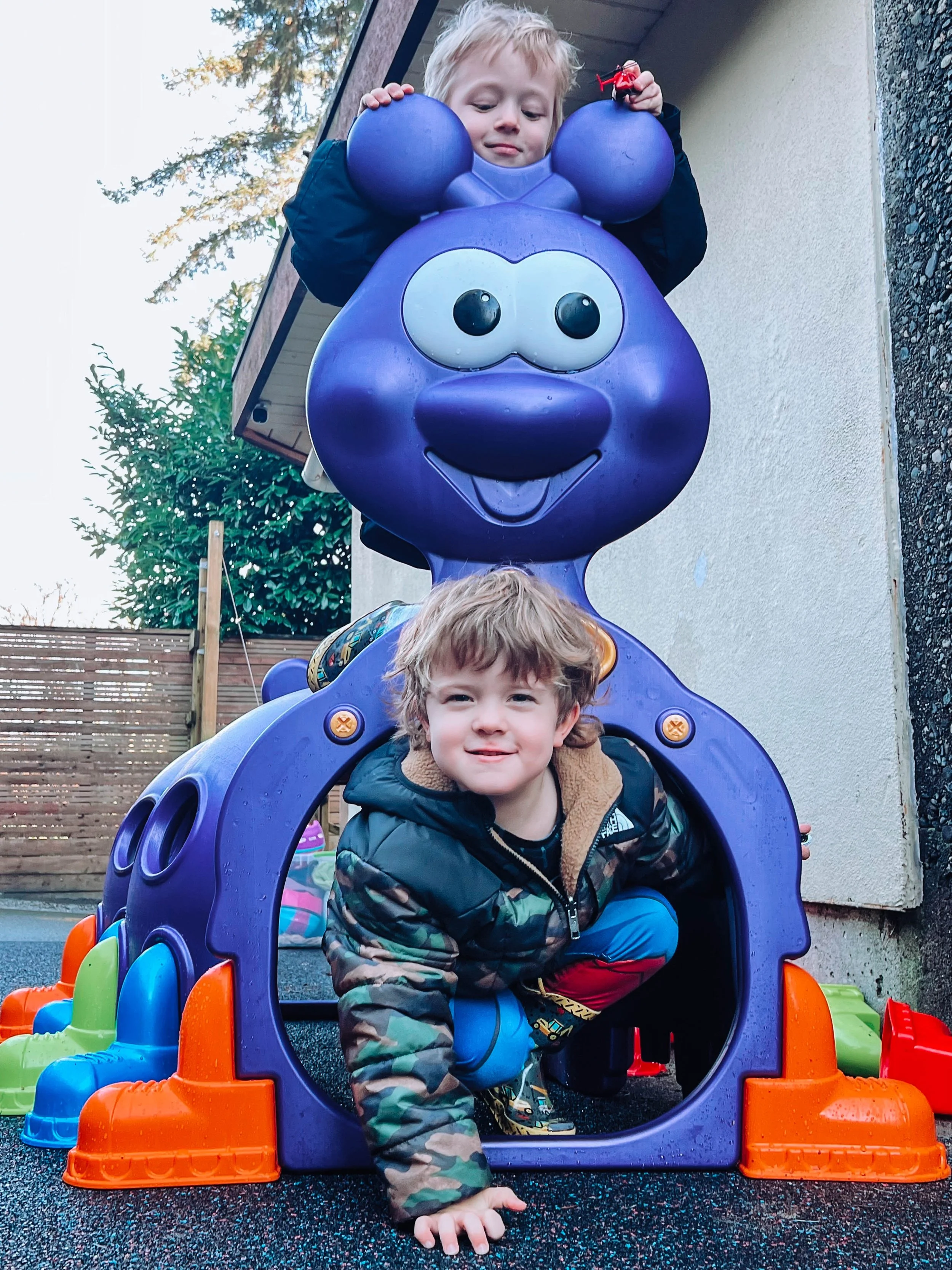Two children playing on a purple Winnie the Pooh play structure outside. The boy is crawling through the opening at the bottom, smiling, while the girl is sitting on top, holding a small red toy and looking down.