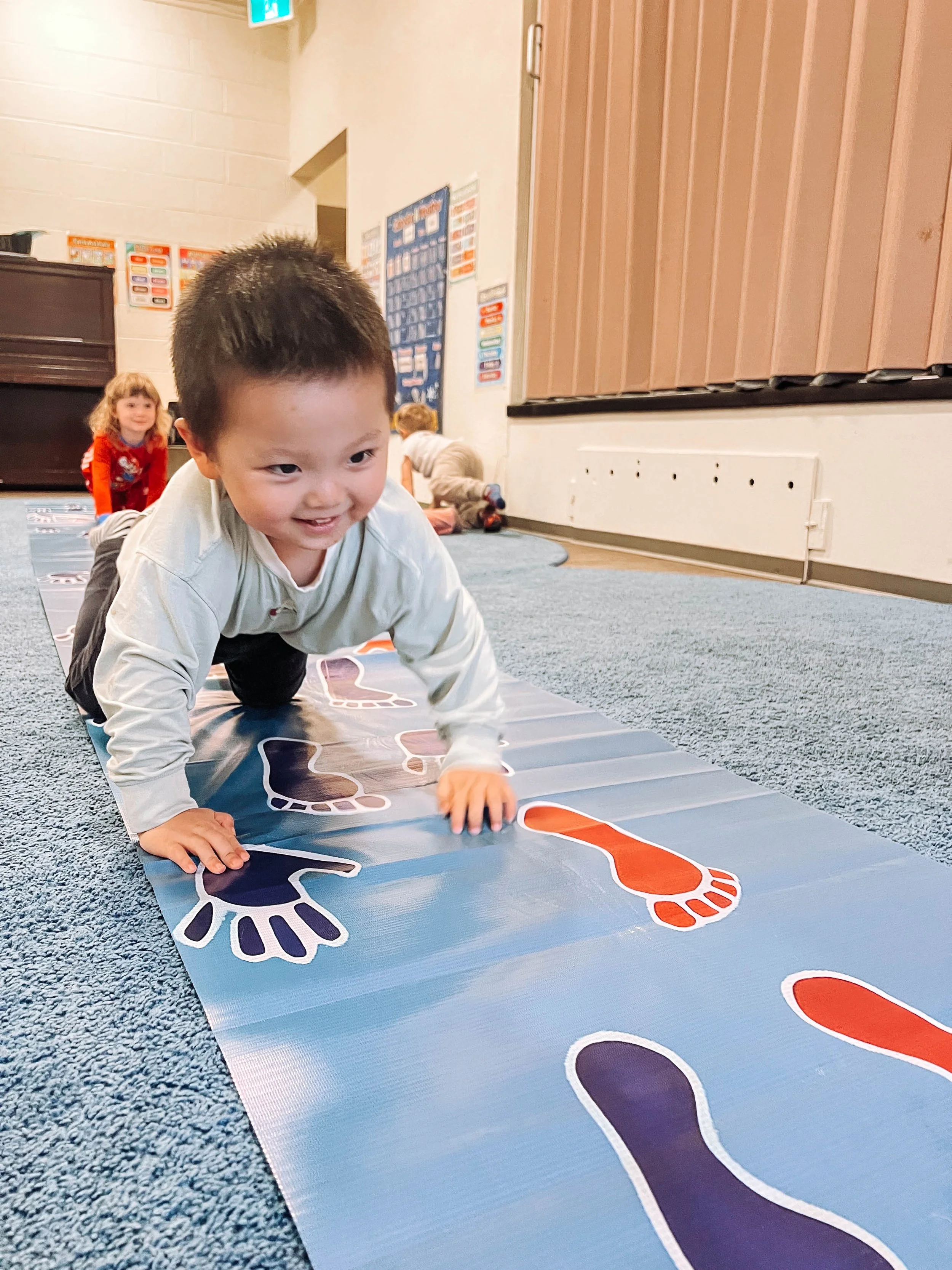 Several large yachts docked at a marina with several smaller boats, a forest, and a building in the background under cloudy skies.
