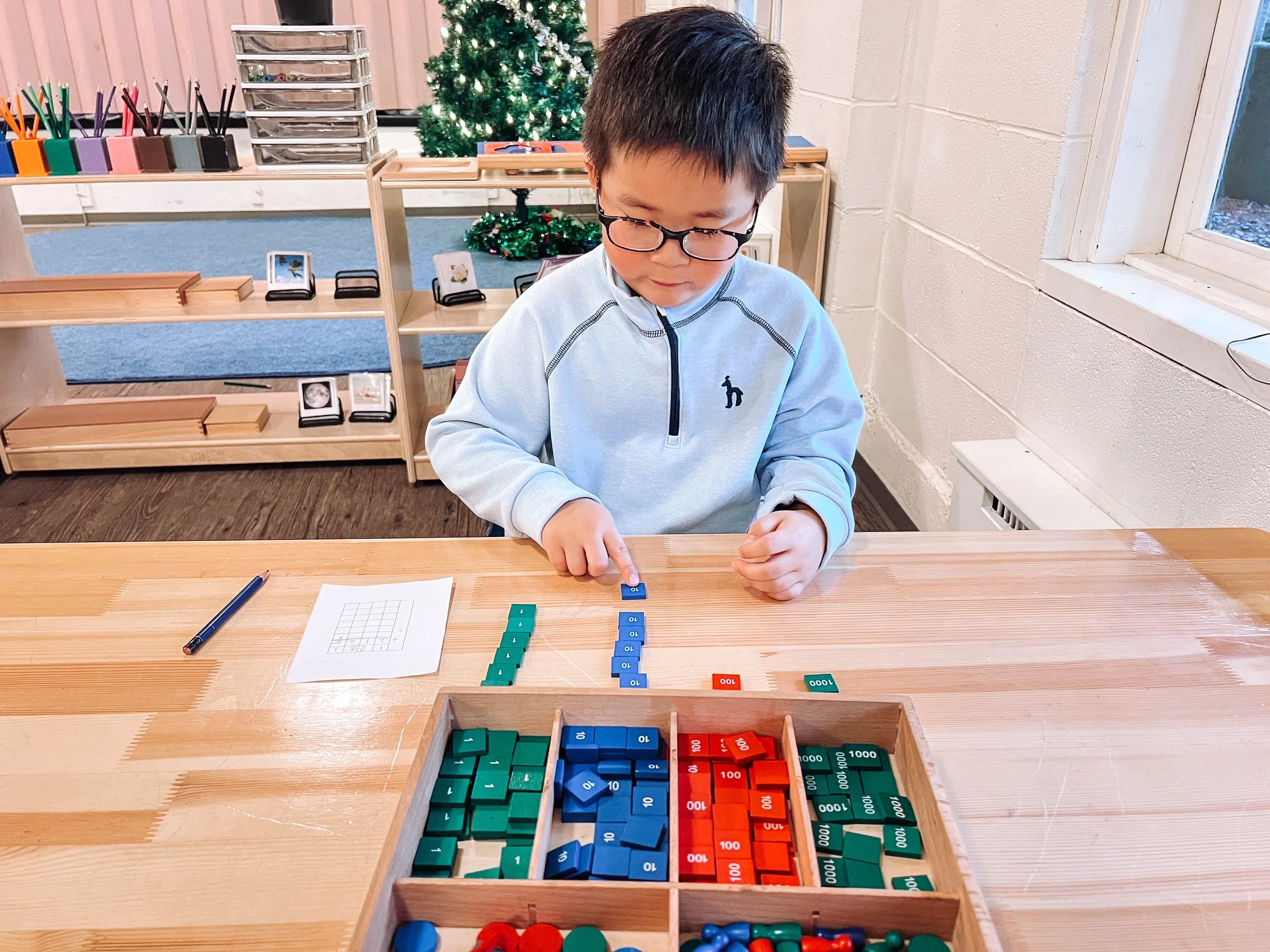 A young boy wearing glasses and a light gray sweatshirt is sitting at a wooden table, playing with colorful counting blocks, with a small sheet of paper and a pencil nearby. In the background, there's a Christmas tree, shelves with various items, and