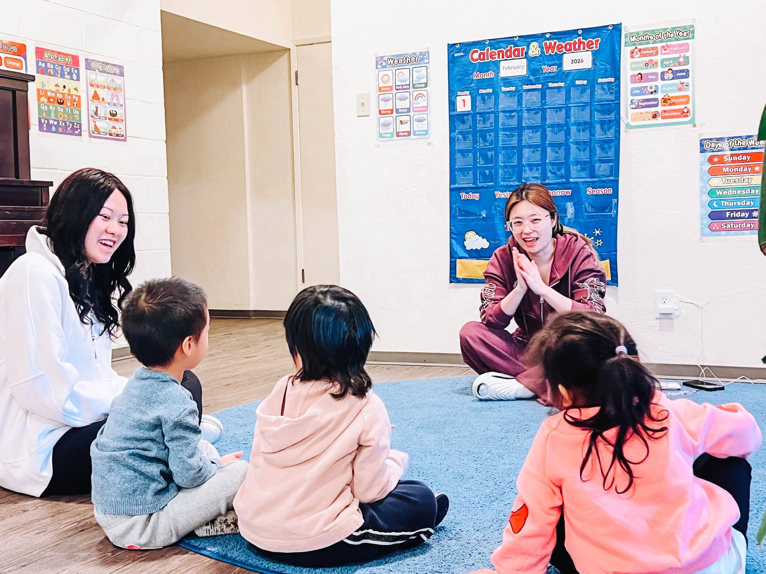 Two women and four children sitting on a blue rug in a classroom, engaging in a storyTime or lesson, with educational posters and a calendar on the wall.