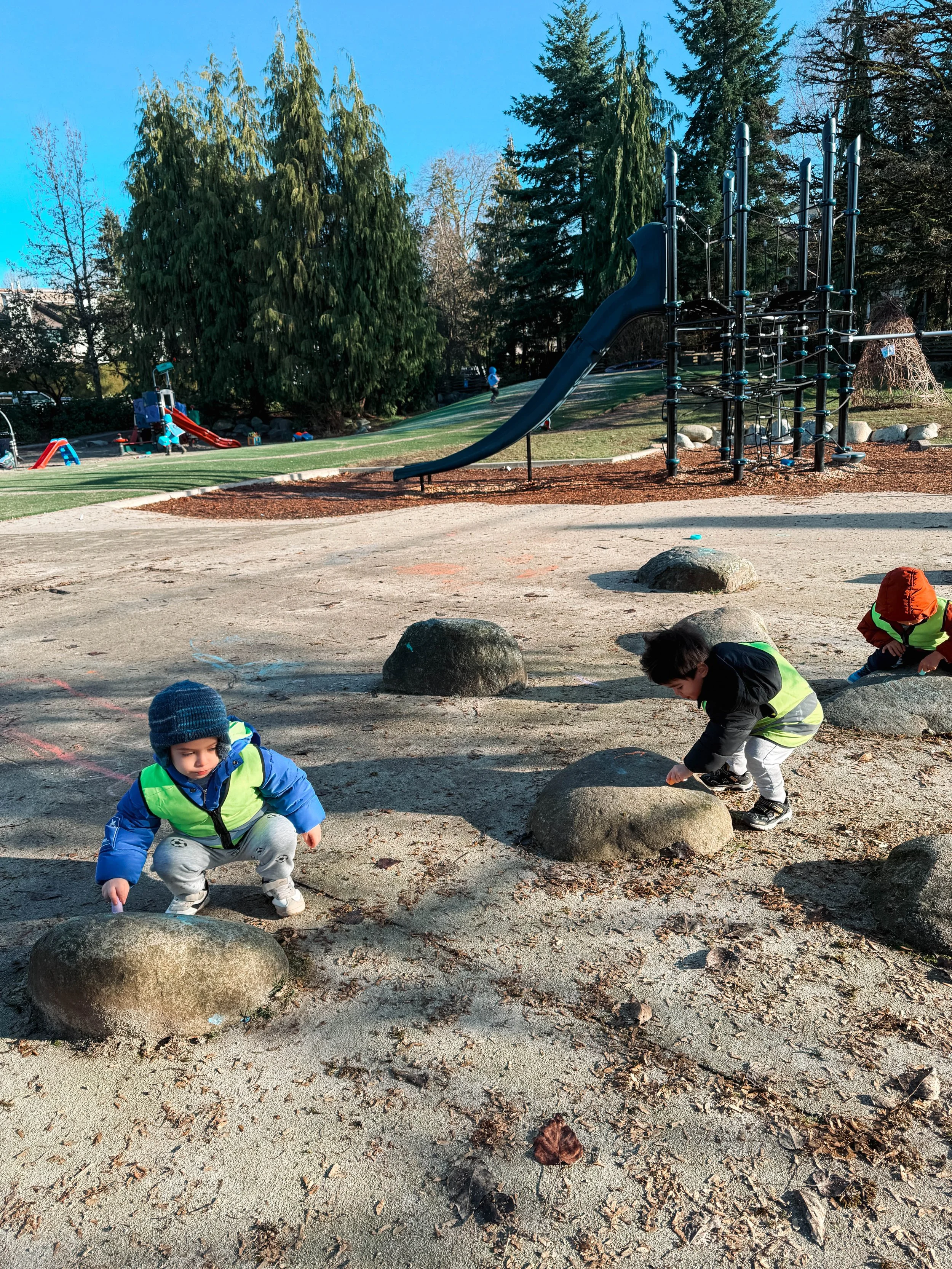 Three children playing on large rocks at a playground, with a slide and climbing structure in the background, surrounded by trees on a sunny day.