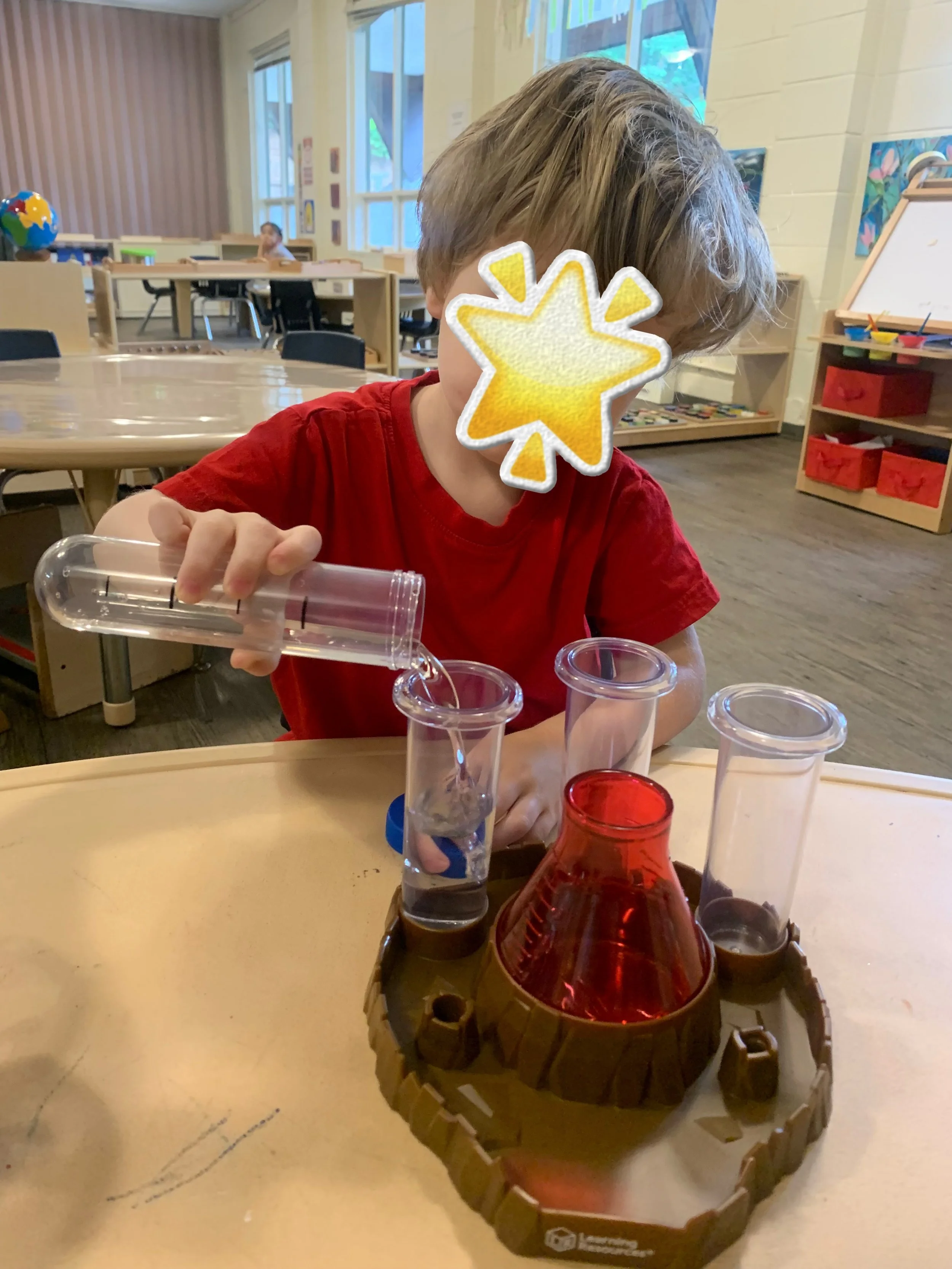 A young boy with a star sticker over his face is conducting a science experiment in a classroom, pouring a clear liquid from a test tube into a graduated cylinder.
