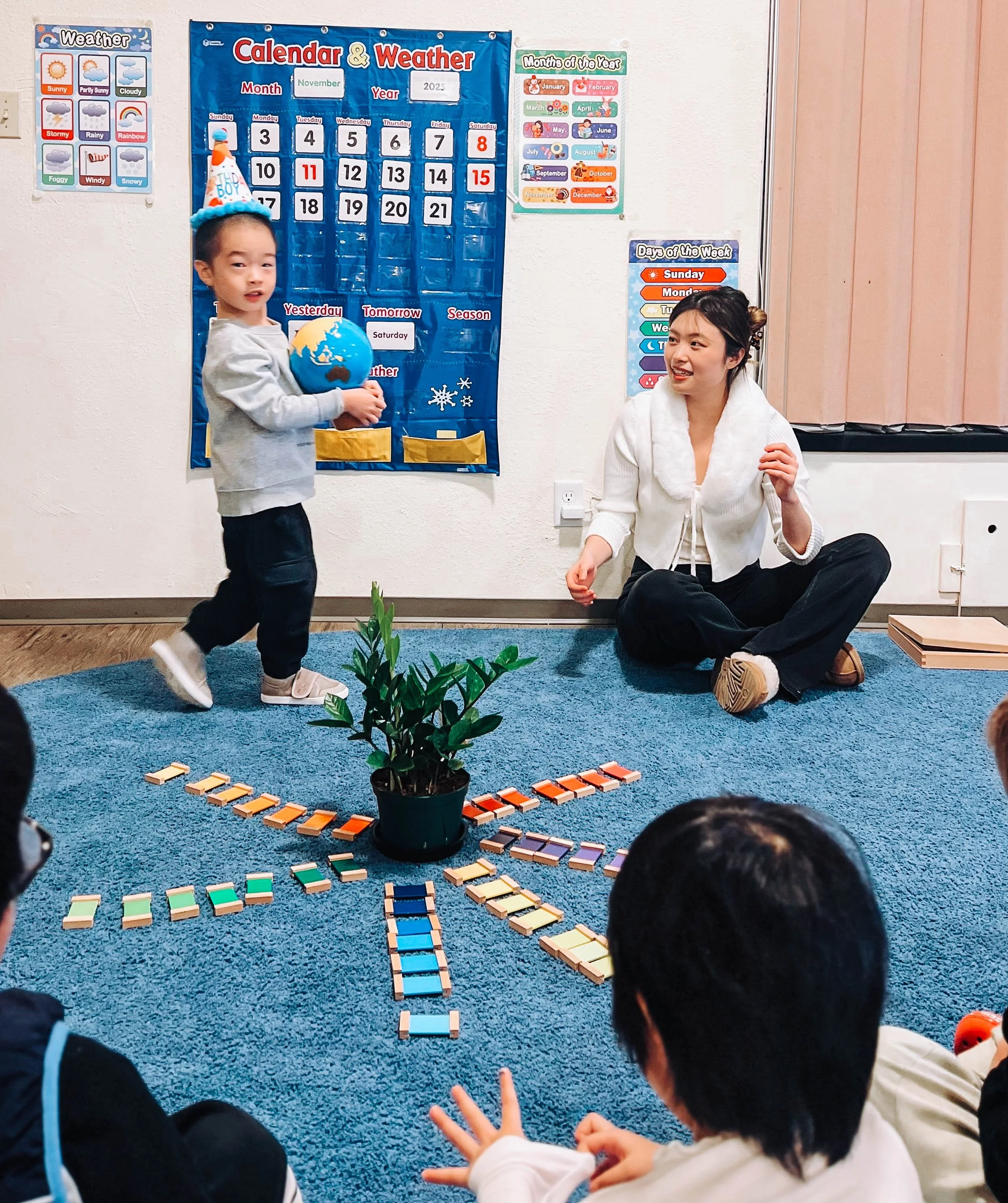 A young child wearing a birthday hat holding a globe, standing in front of a group of children seated on a blue carpet, with a woman sitting on the floor nearby. The setting appears to be a classroom with educational posters about weather and days of