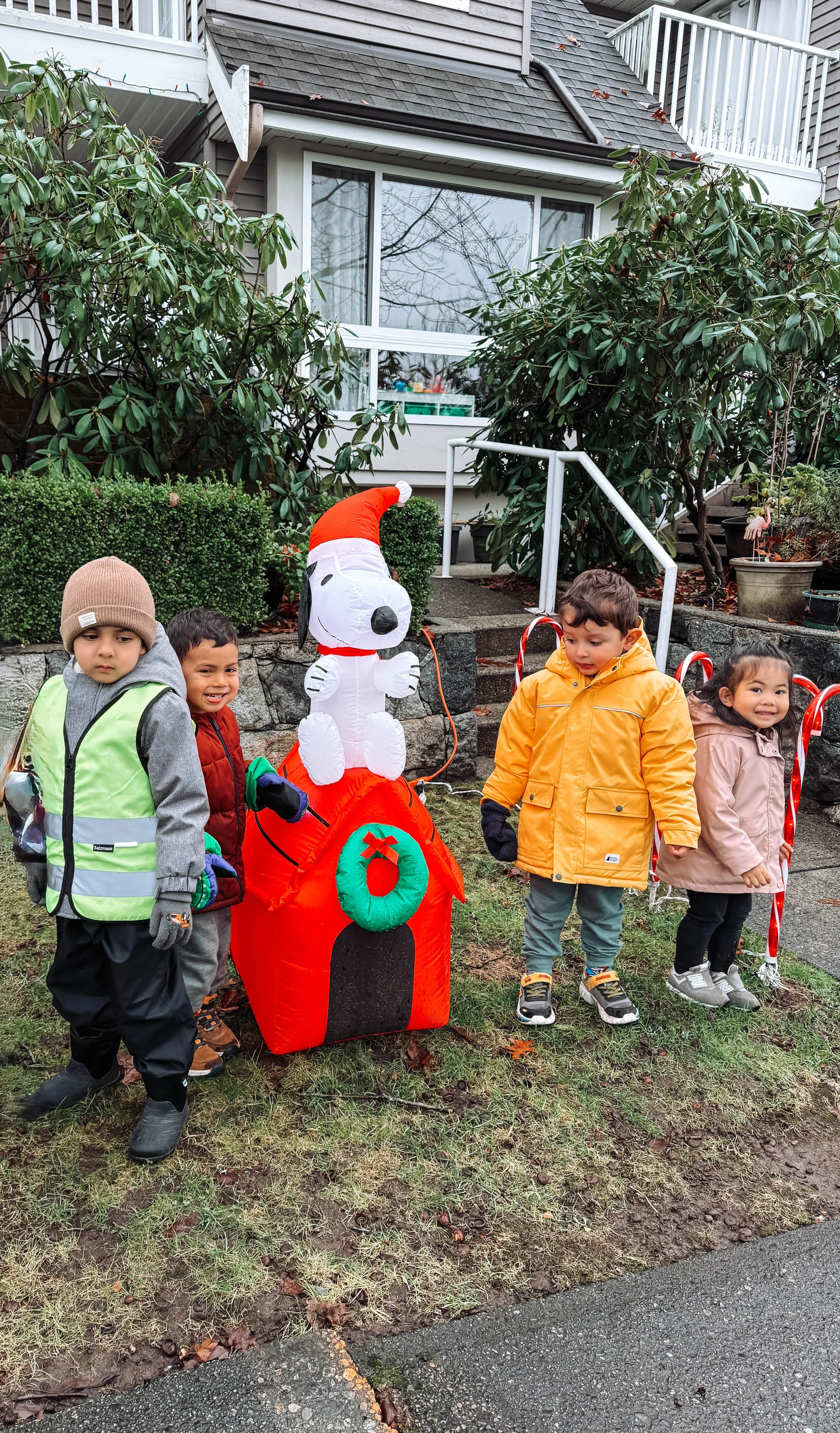 Children dressed in warm clothing standing outdoors next to a large inflatable Santa Claus decoration, with a house and bushes in the background.
