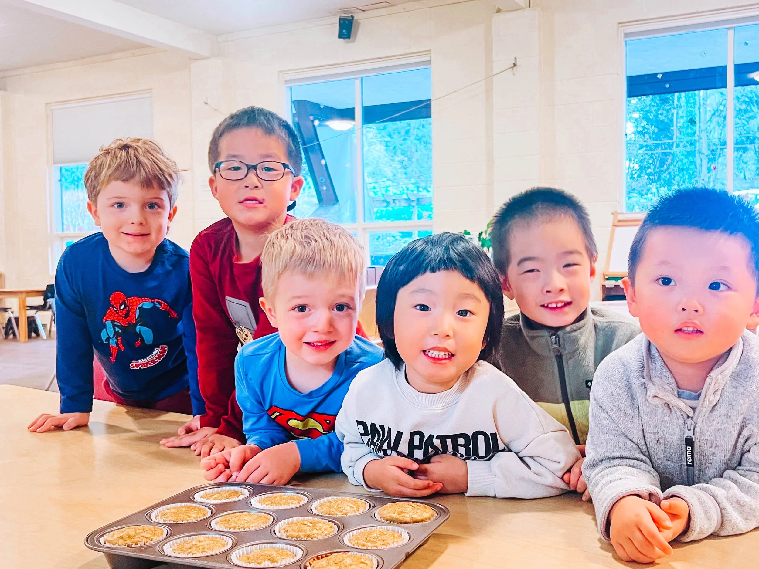A group of six young children leaning on a table with a tray of baked cupcakes, in a brightly lit room with large windows.