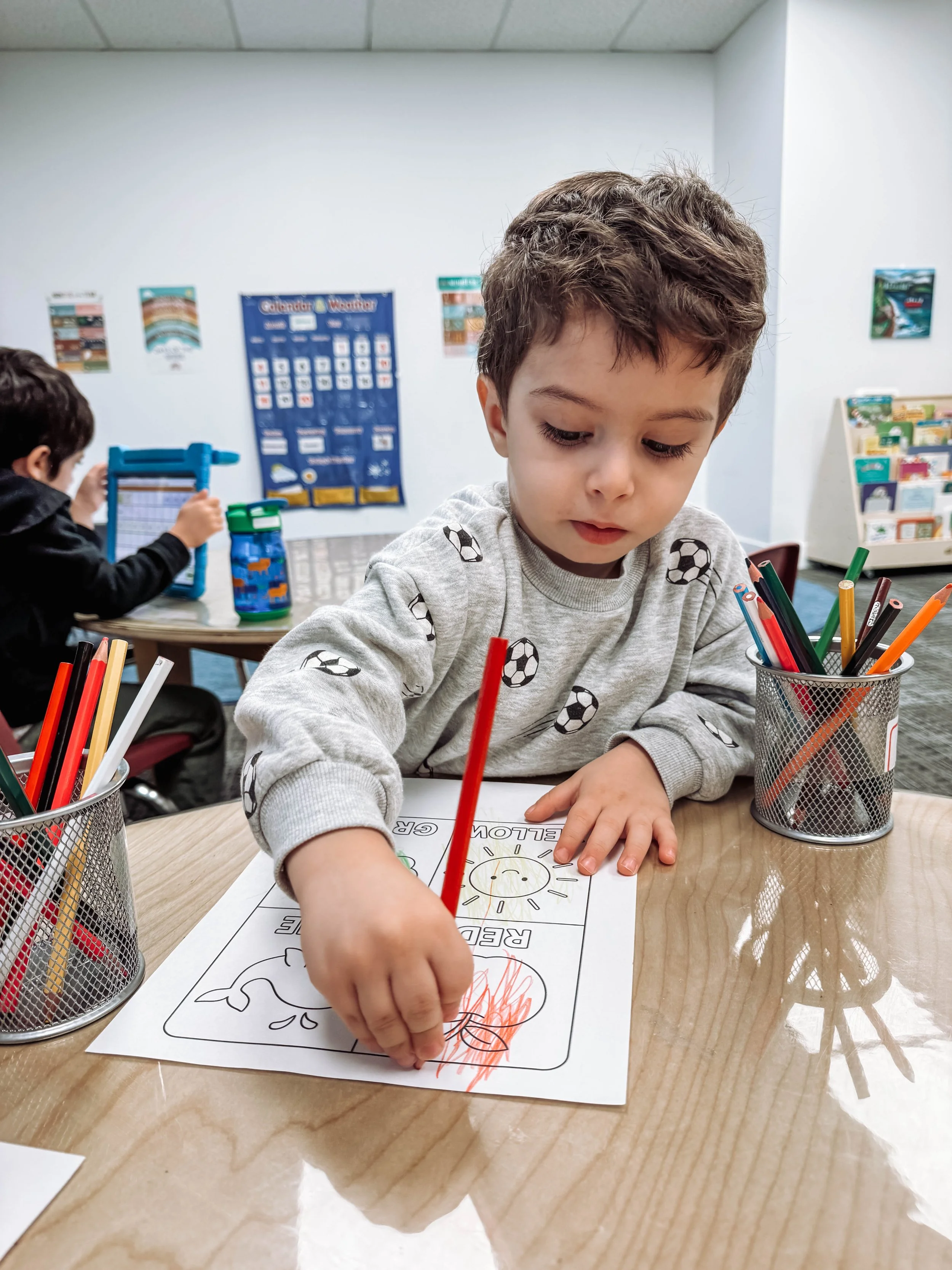 A young boy coloring a worksheet at a classroom table with colored pencils. Another child is visible in the background working at a different table.