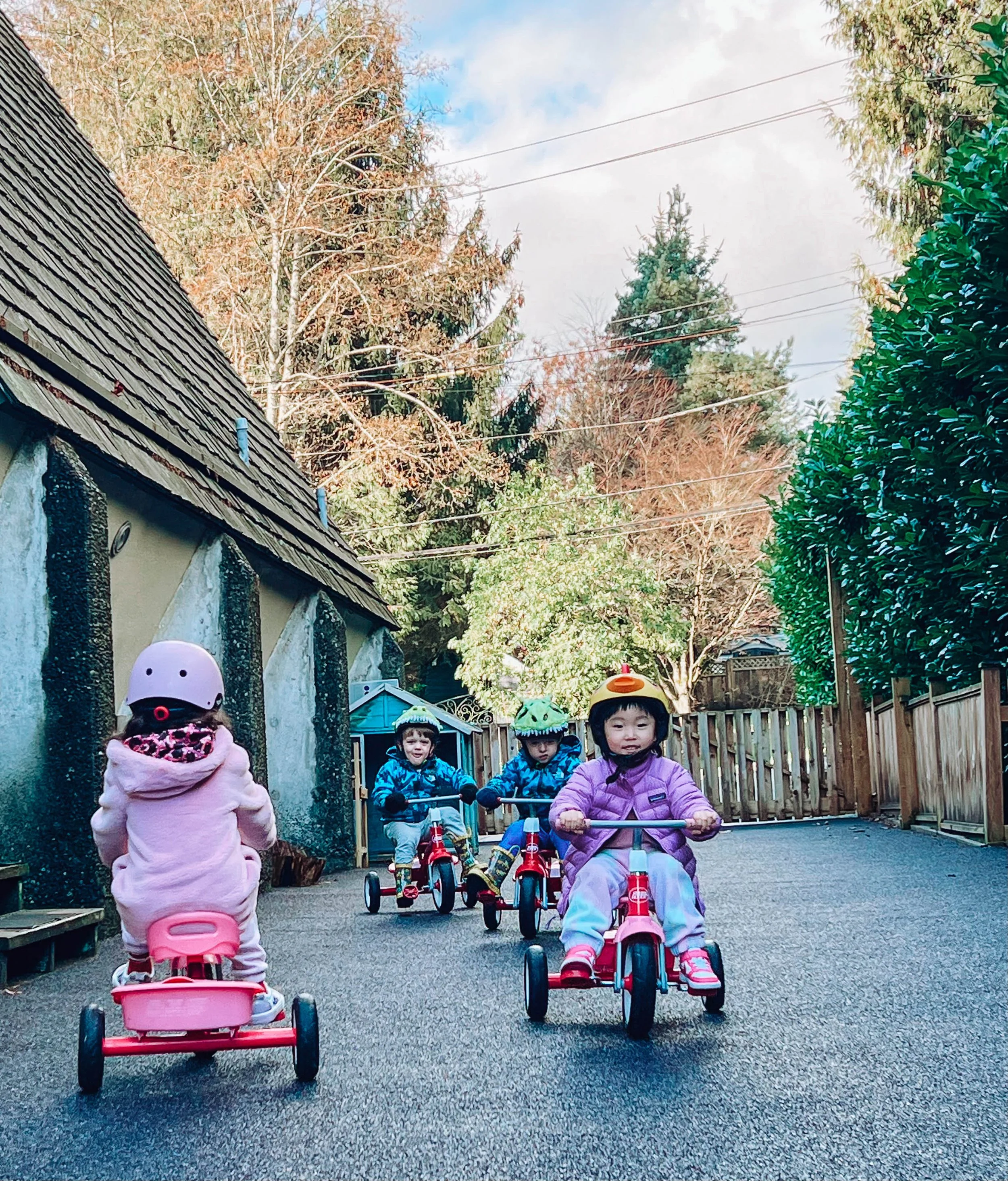 Four young children riding tricycles outside on a cleared driveway. Three children are wearing helmets, and one is dressed in a dinosaur costume. The setting has trees and a wooden fence, with a cloudy sky overhead.