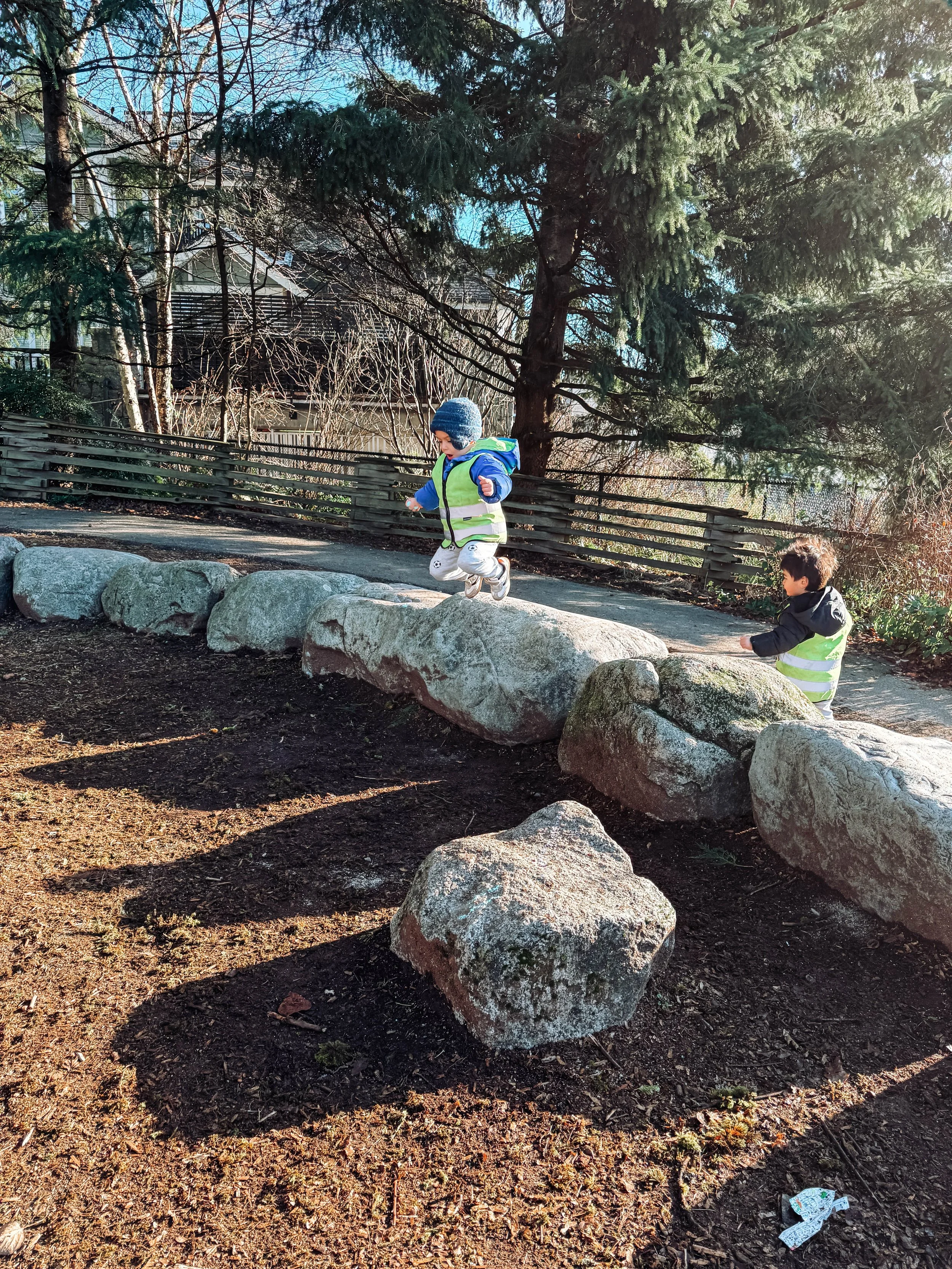 Two young children wearing reflective vests playing on large rocks outdoors in a park.