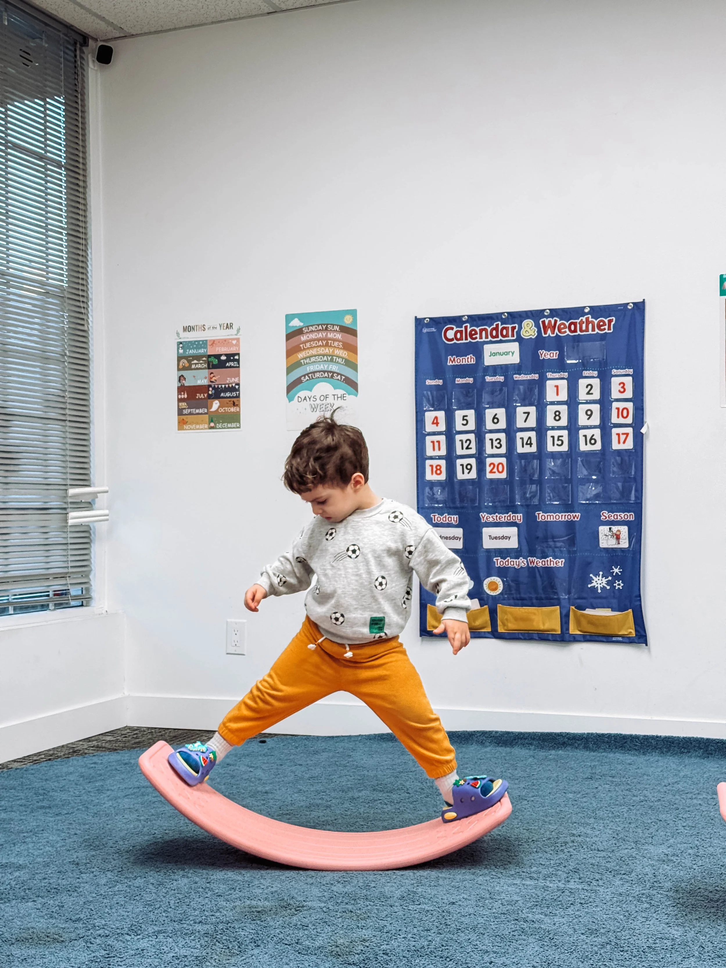 Young boy balancing on a pink curved balance board in a classroom with educational posters and a calendar on the wall.