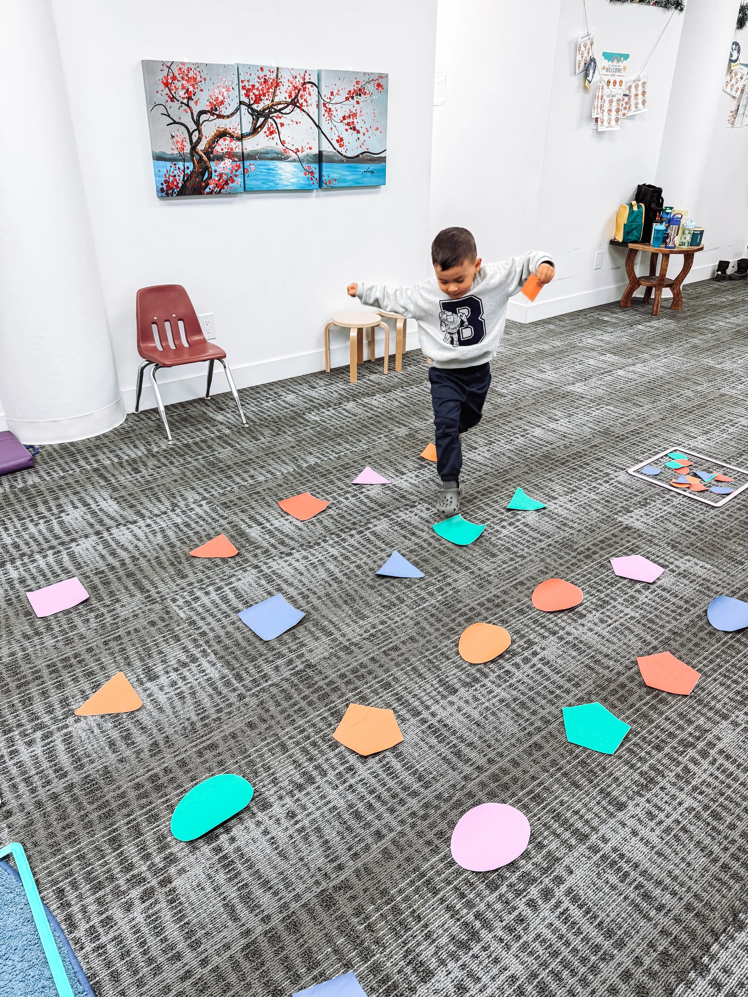A young boy wearing a gray sweatshirt with the letter 'B' on it and black pants is jumping over colorful paper shapes on the carpeted floor of a room. The room has white walls, a red chair, small stacked stools, and a wall art piece depicting a tree 