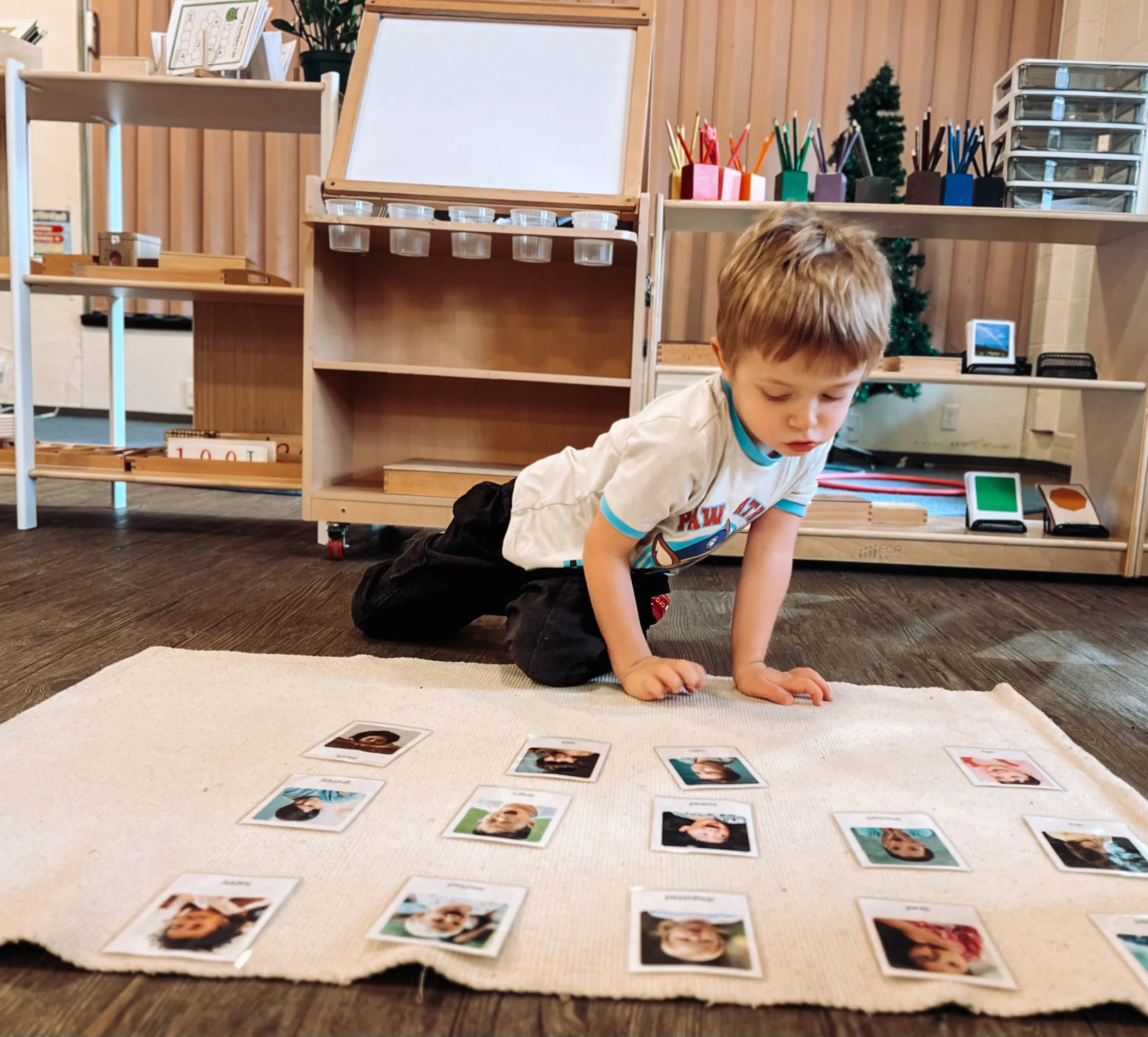Young boy playing a matching game with photo cards on a cream-colored rug in a classroom or activity room.