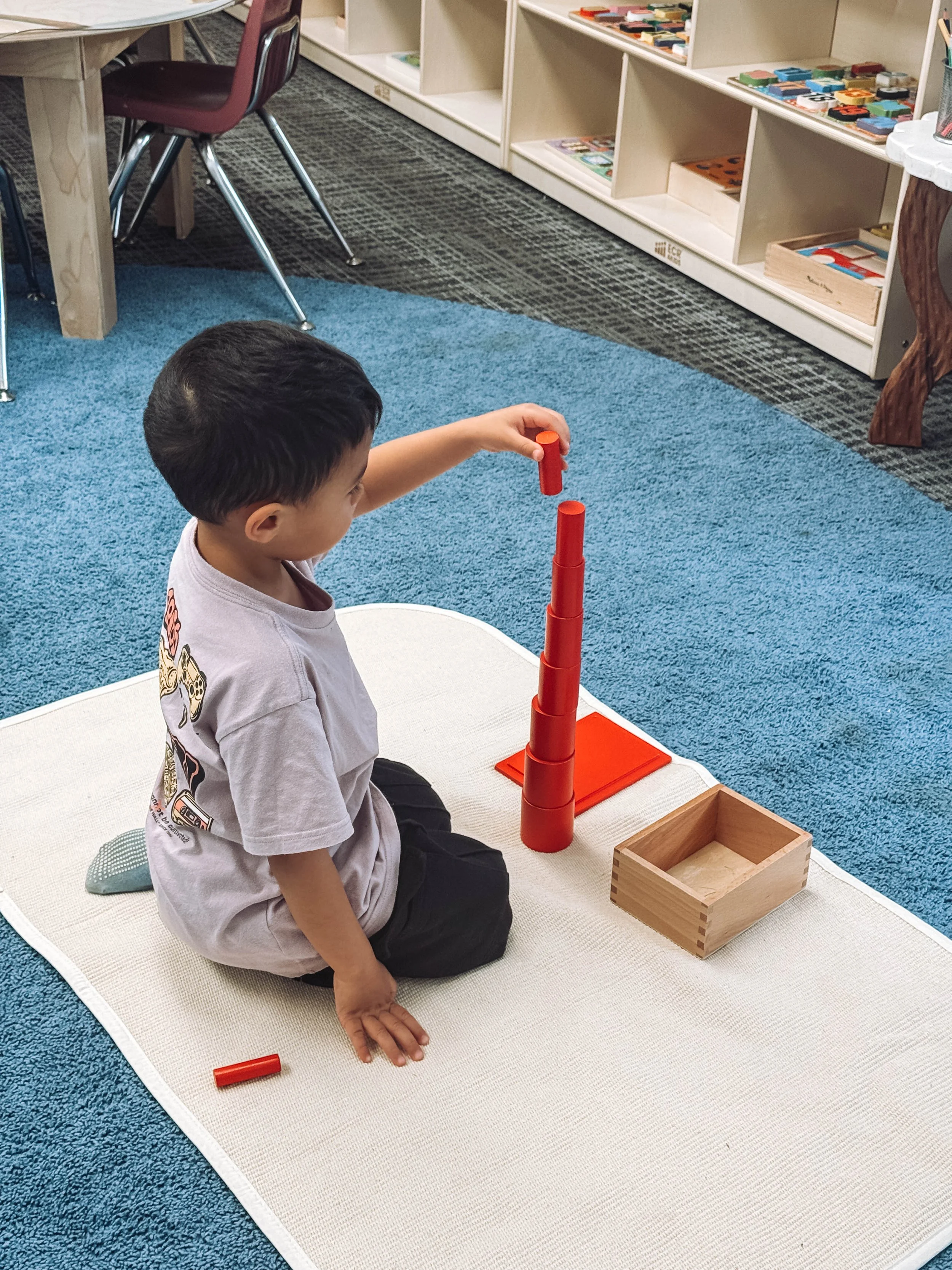 A young boy sits on a white mat in a classroom, stacking orange cylindrical blocks into a tower. There is a wooden box and a small flat orange board nearby, with shelves filled with educational toys and books in the background.