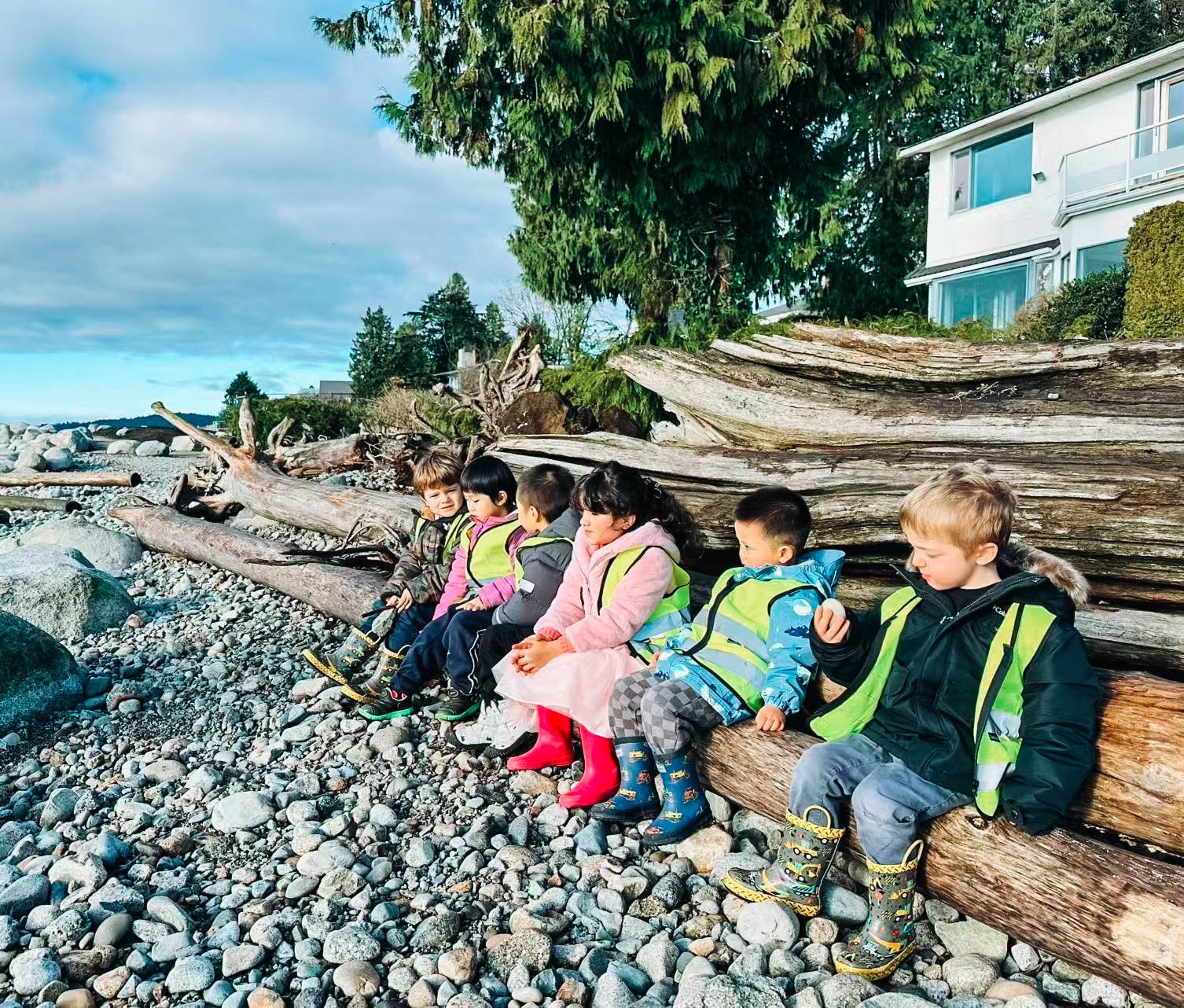 Six children sitting on logs and rocks along a rocky beach, wearing colorful jackets and rain boots.