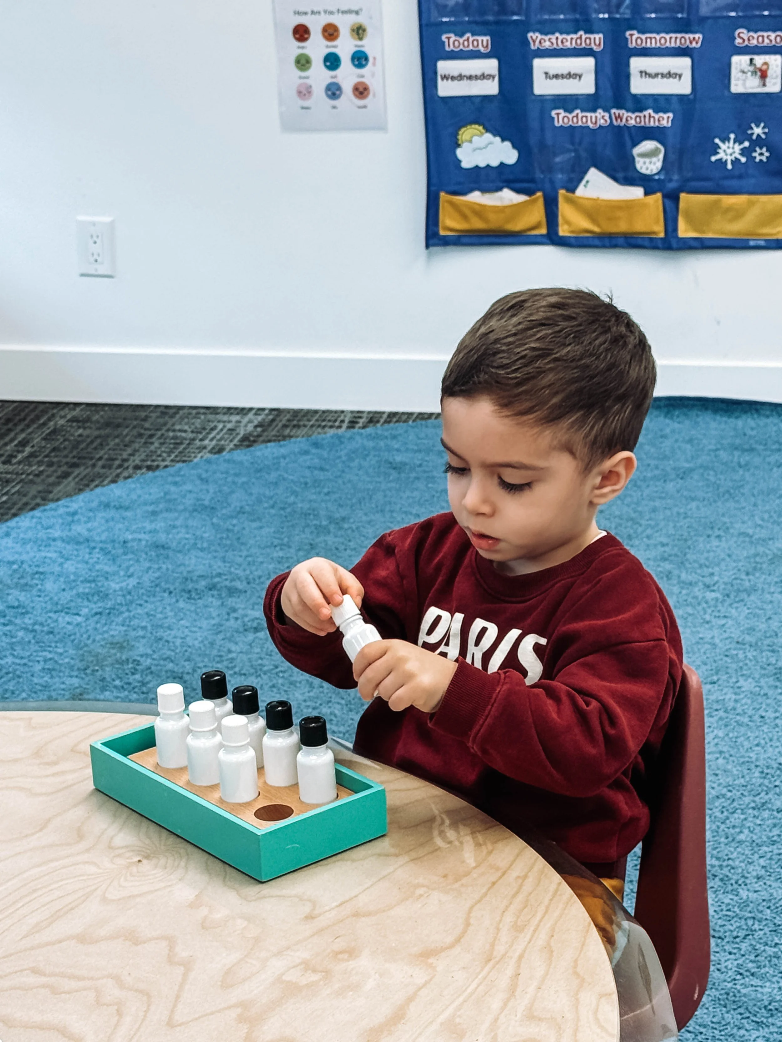 A young boy in a red sweatshirt with the word 'Paris' on it, sitting at a round wooden table in a classroom, is playing a game with small white bottles with black caps, which are arranged in a neat row inside a teal tray.