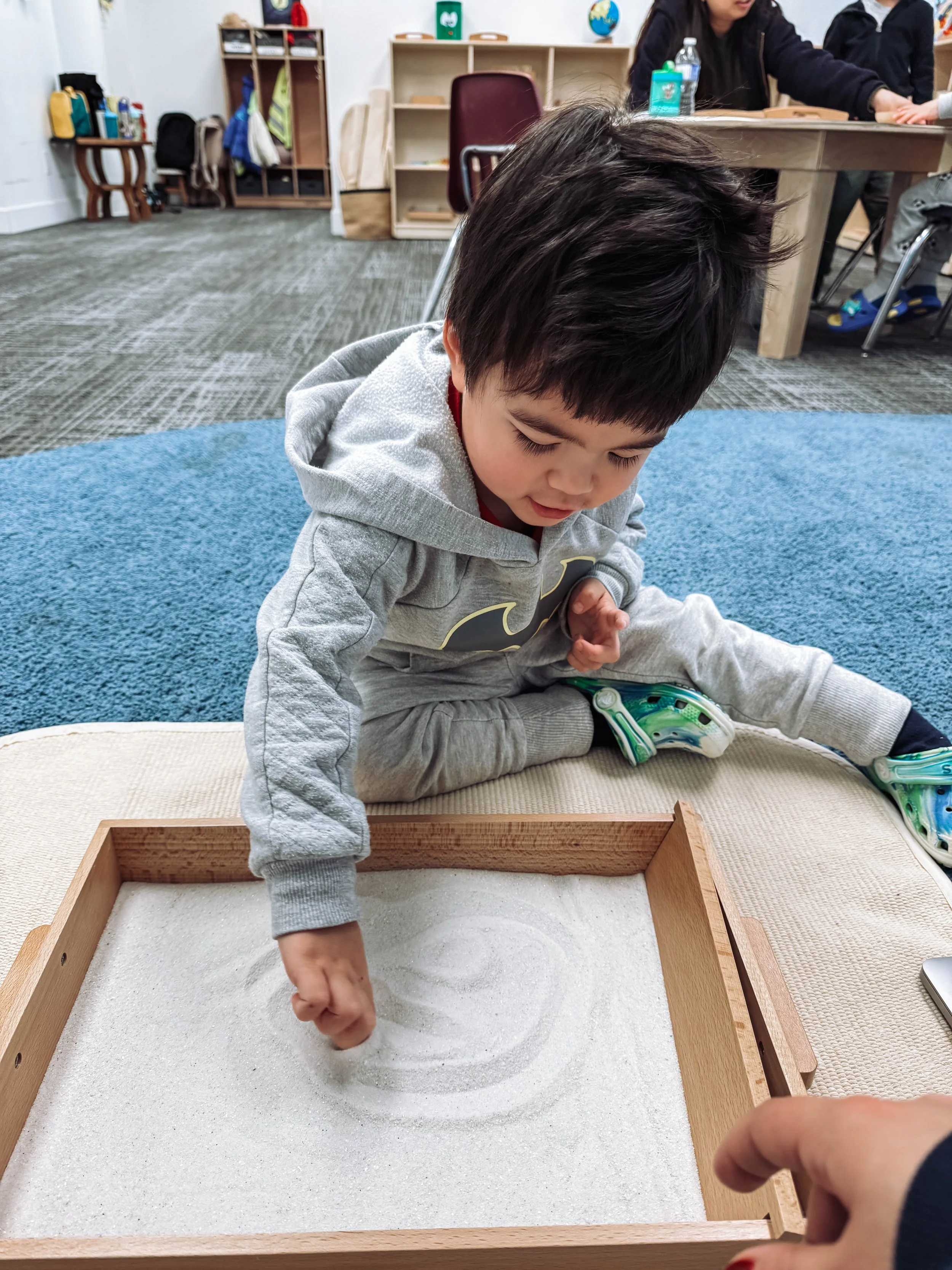 A young boy dressed in gray hoodie and sweatpants sitting on a cushioned surface, using his finger to draw in a tray filled with white sand in a classroom setting.