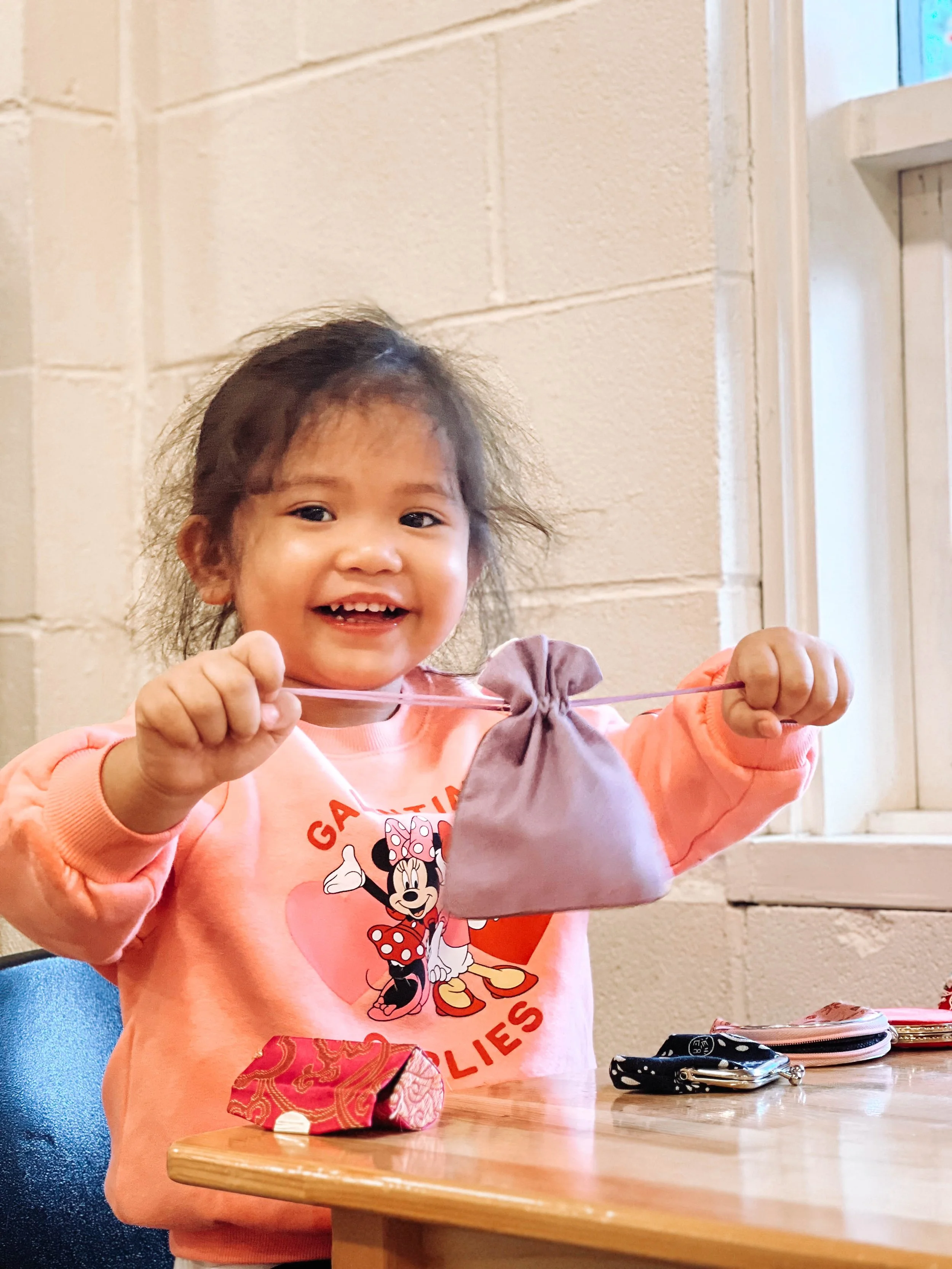 A young girl smiling and holding a small gray drawstring bag on a string in a room with beige brick walls, sitting at a wooden table with various small items.