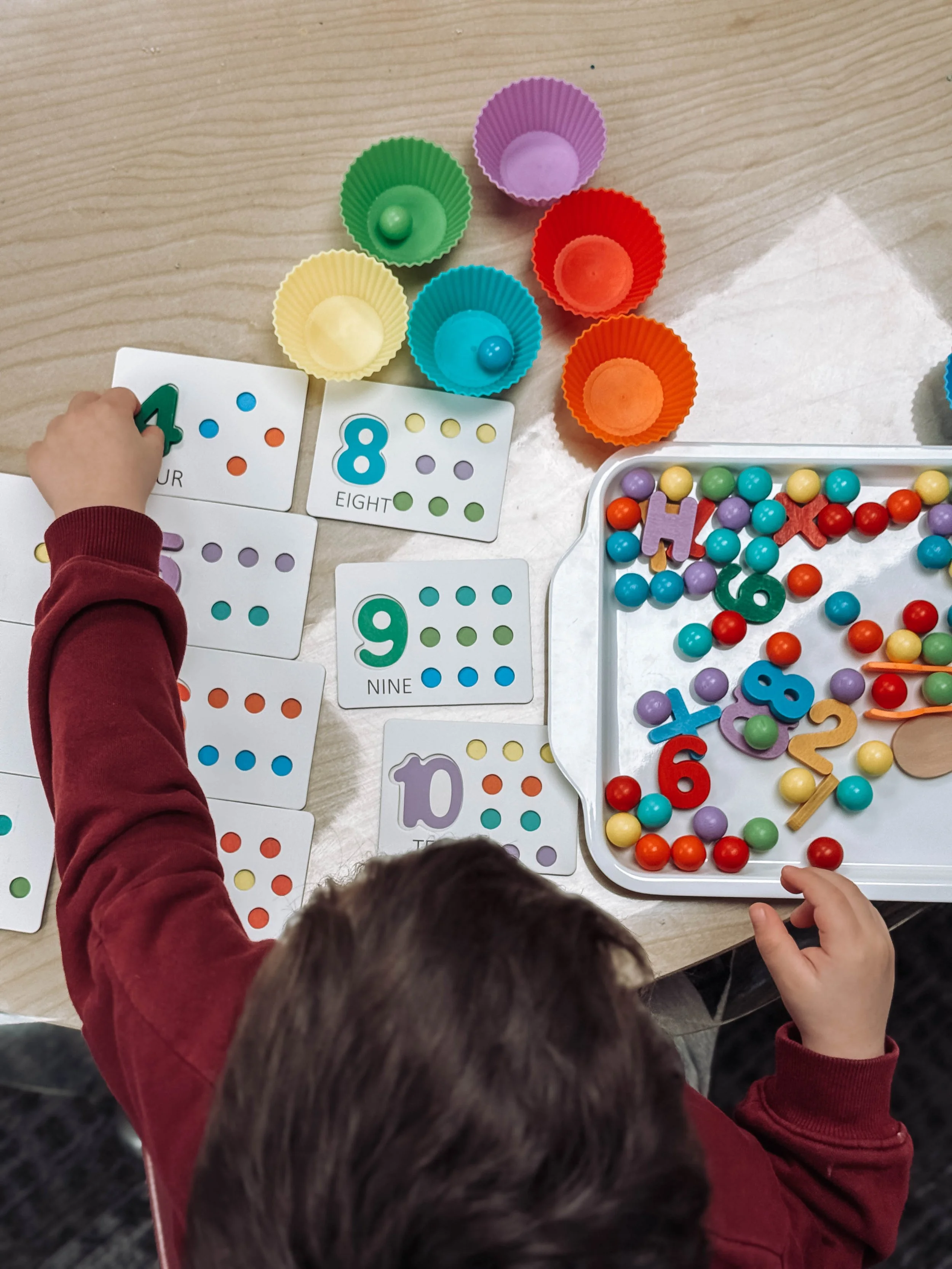 Child in a red long-sleeve shirt playing with colorful teaching materials, including number cards, dot count cards, and a tray of multicolored beads and foam shapes.
