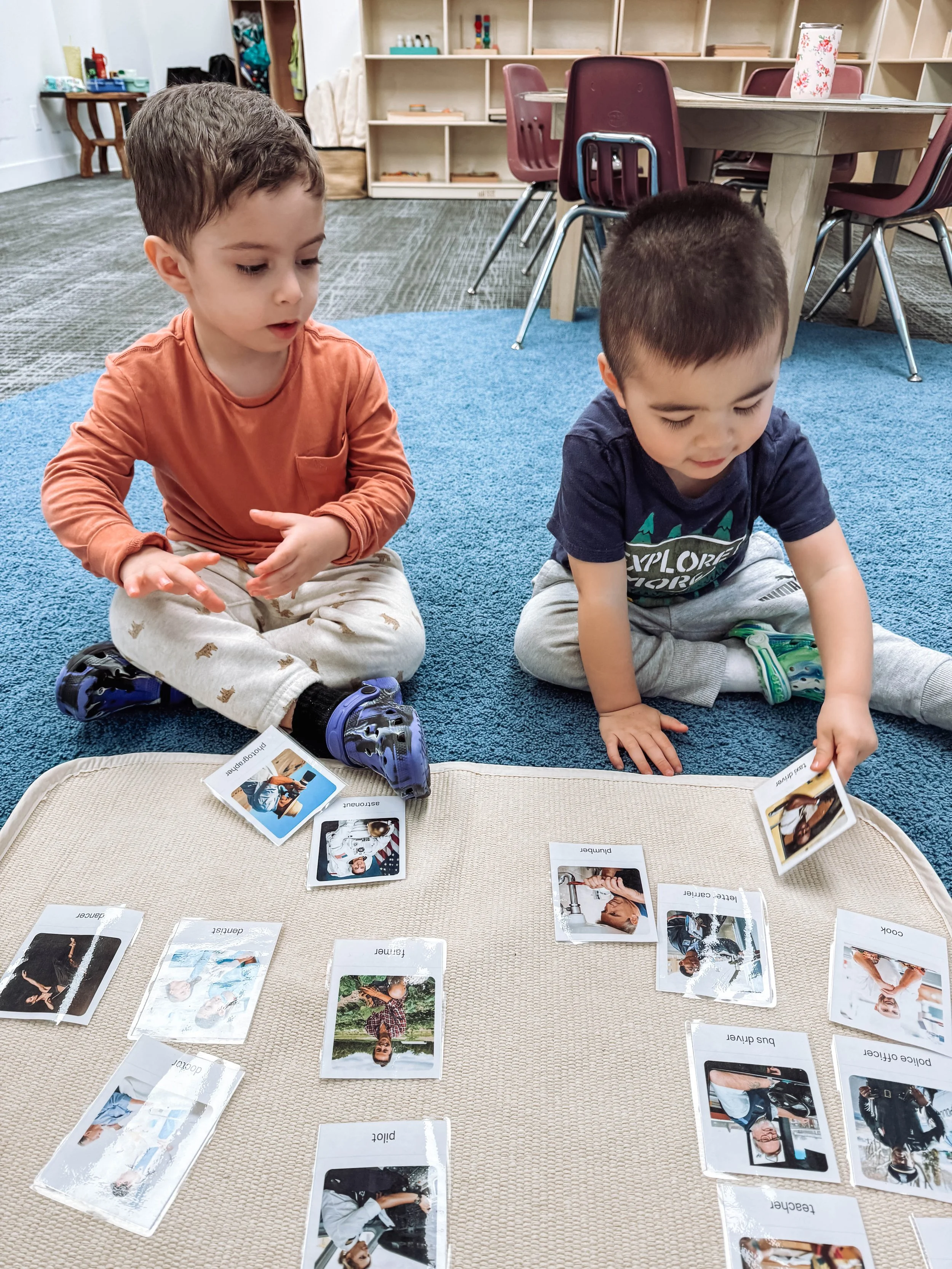 Two young boys playing a matching game with picture cards on a beige mat in a classroom.