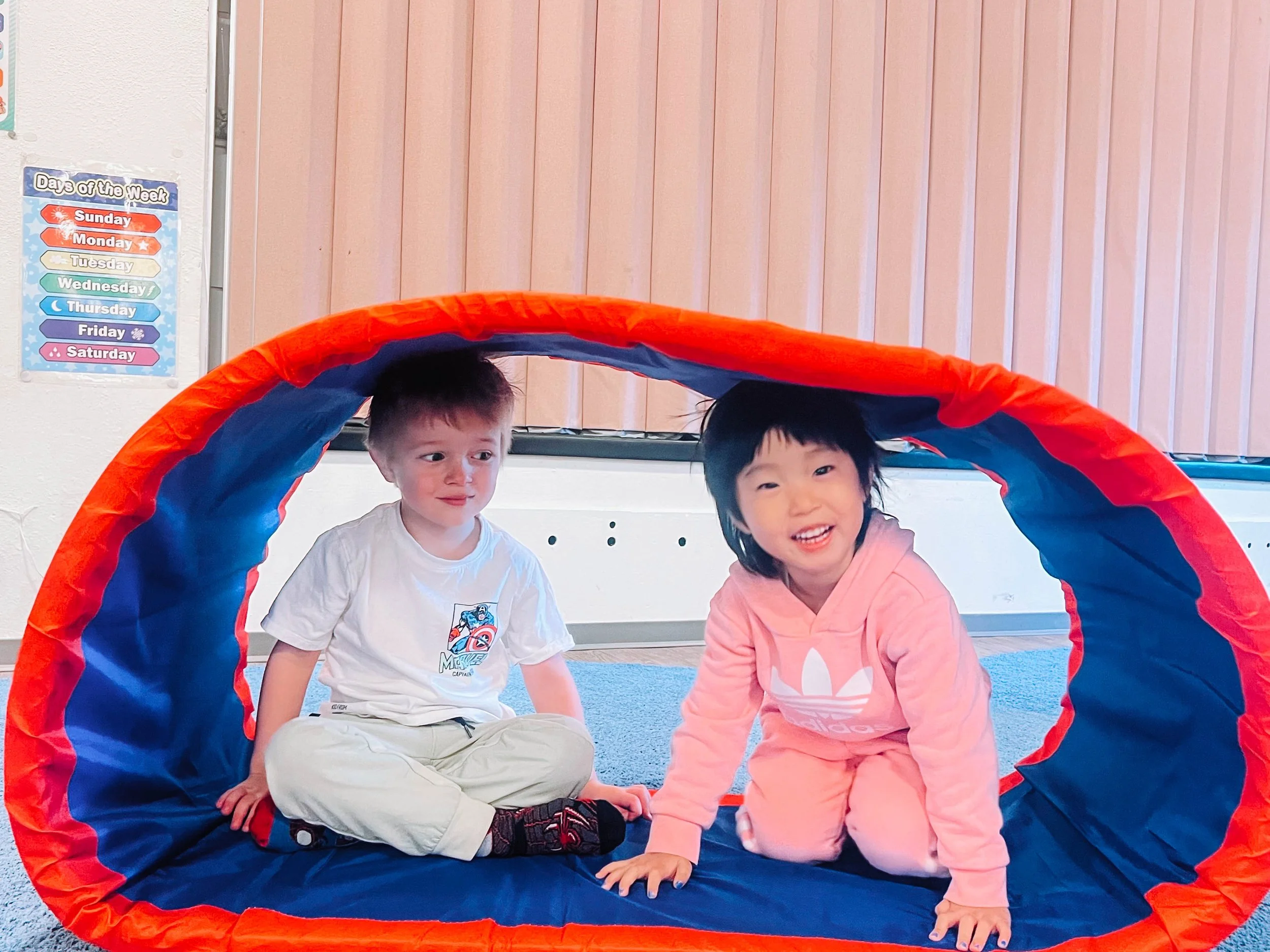Two children playing inside a red and blue fabric tunnel at a classroom setting.