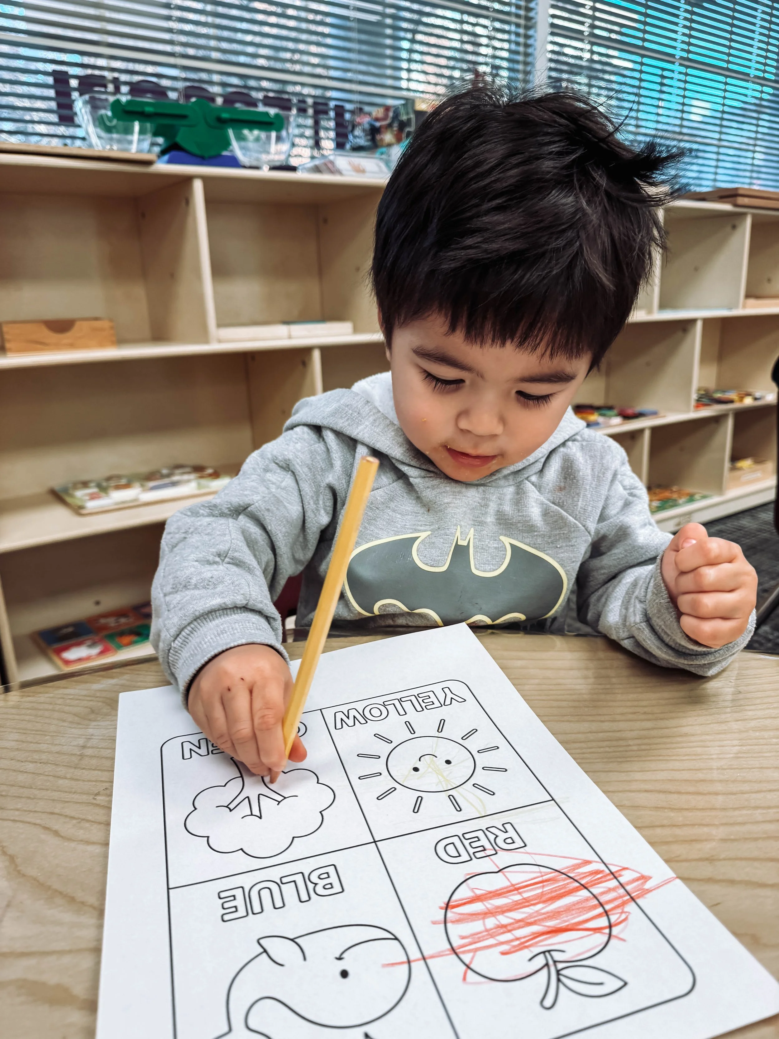 A young boy coloring a weather-themed worksheet with a crayon in a classroom.