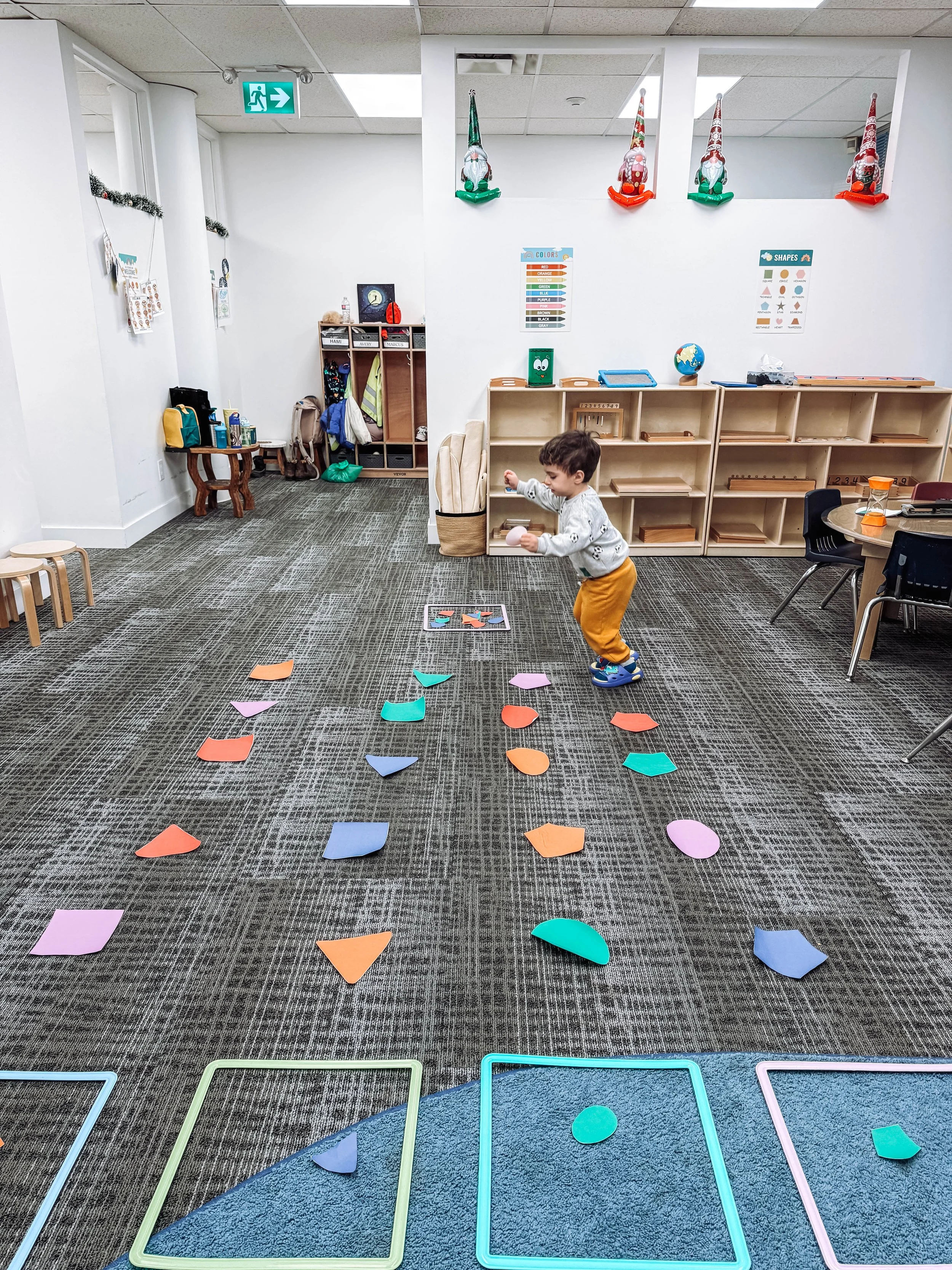 A young boy is playing with colorful paper shapes on a carpeted classroom floor, with open shelving and educational posters in the background.