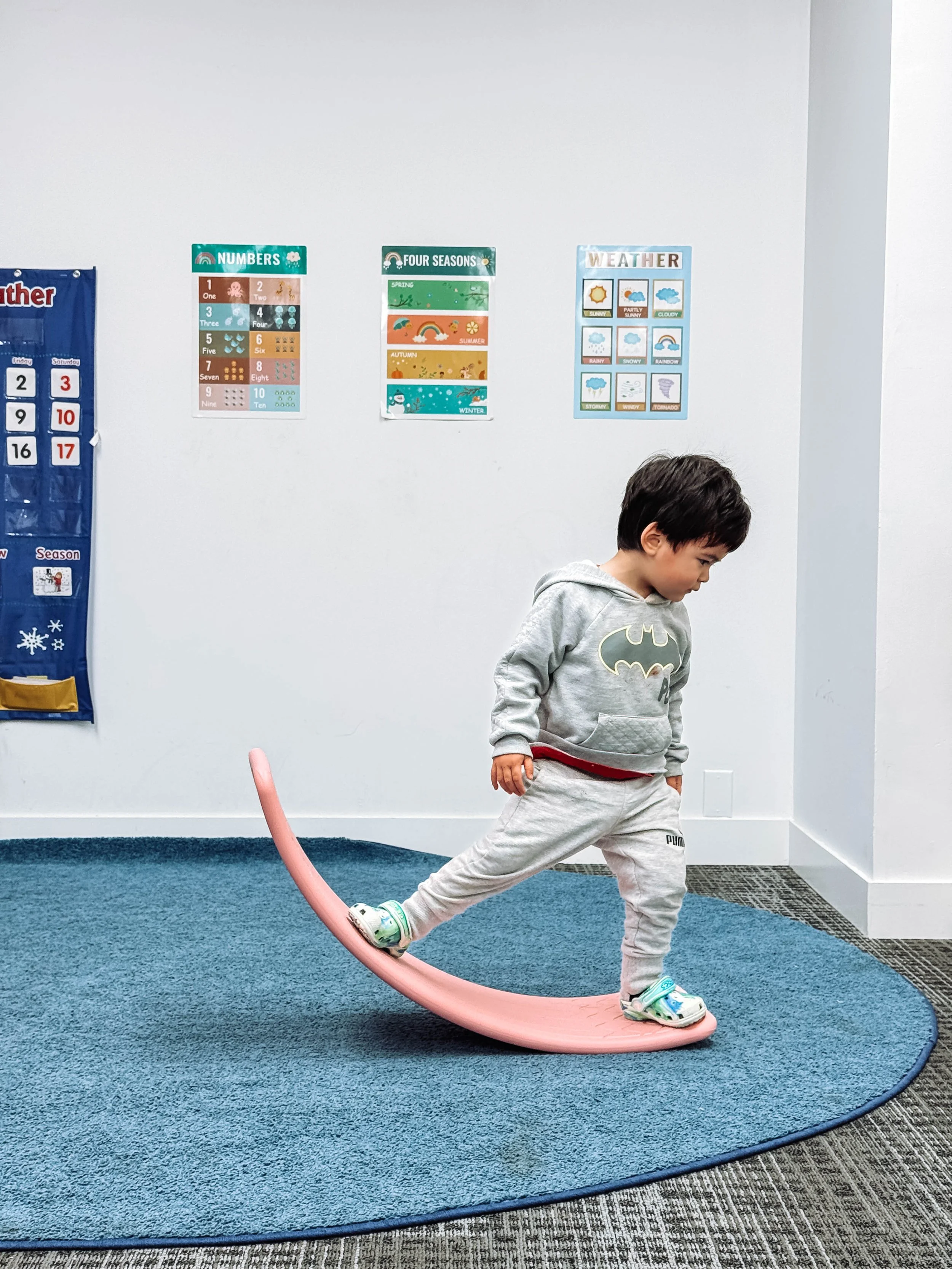 A young boy wearing a gray Batman hoodie and gray sweatpants balancing on a pink curved balance board inside a room with educational posters and a blue carpet.