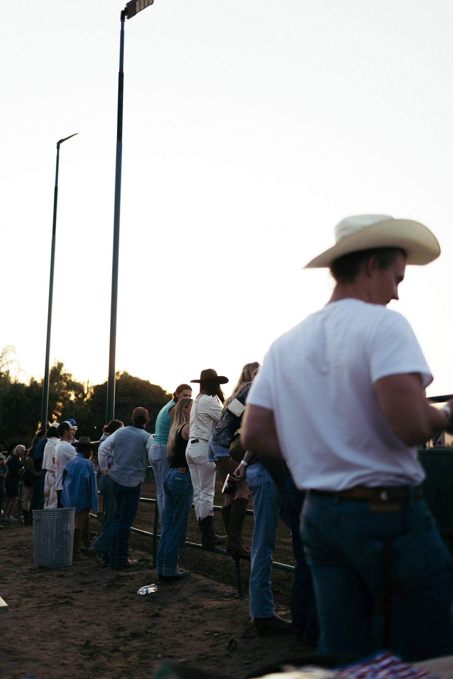 A group of people wearing cowboy hats and casual clothing stands near a fence at an outdoor event, possibly a rodeo, during the day.