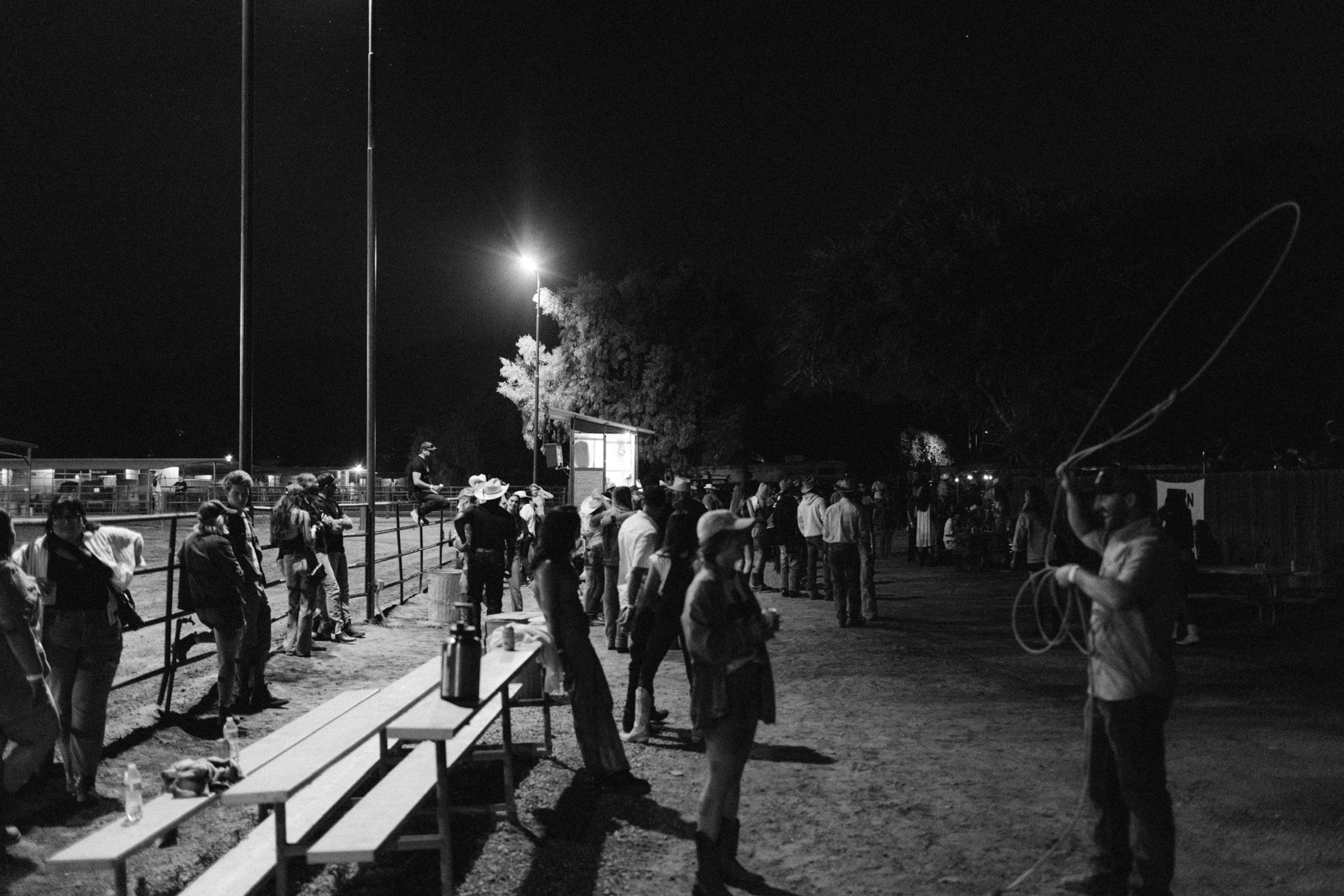Nighttime outdoor gathering with people in cowboy attire, one person practicing lassoing.