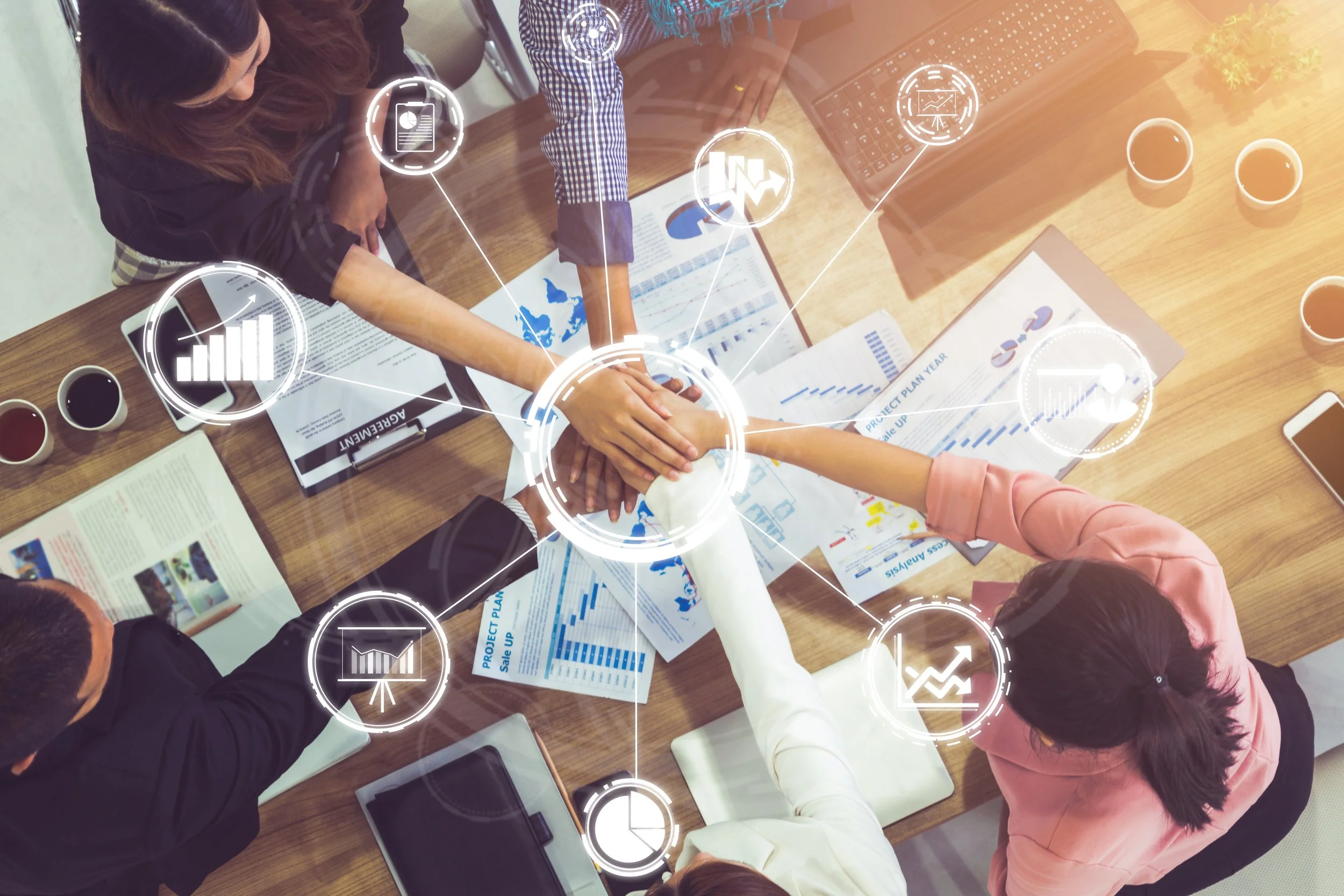 A diverse group of five business professionals sitting around a wooden conference table, engaging in a team huddle with their hands stacked together, surrounded by documents, laptops, and coffee cups. Overlaid digital icons represent data analytics, growth, charts, and graphs.