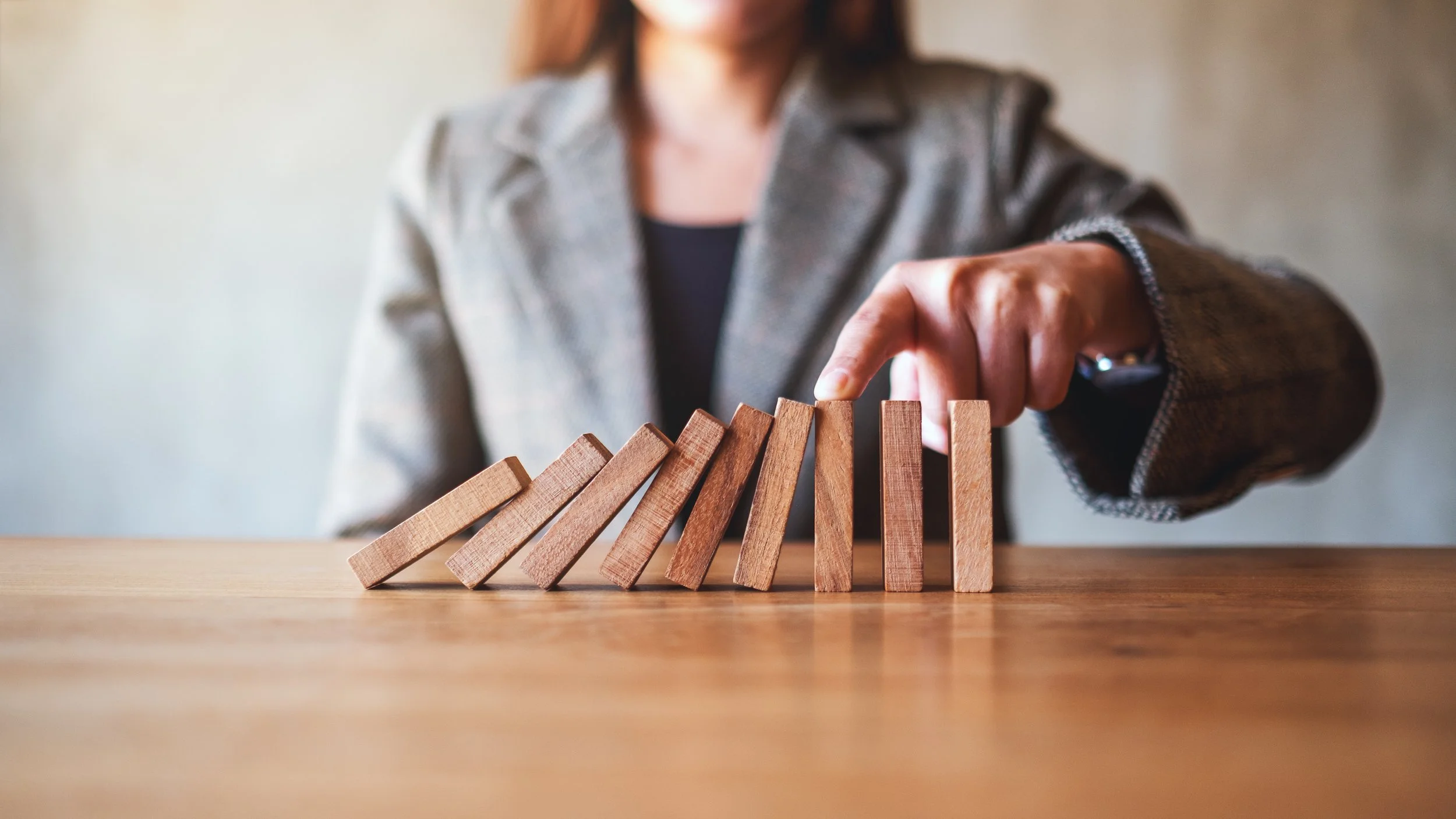 A woman in a blazer stops a row of wooden dominoes from falling by touching the last one with her finger.