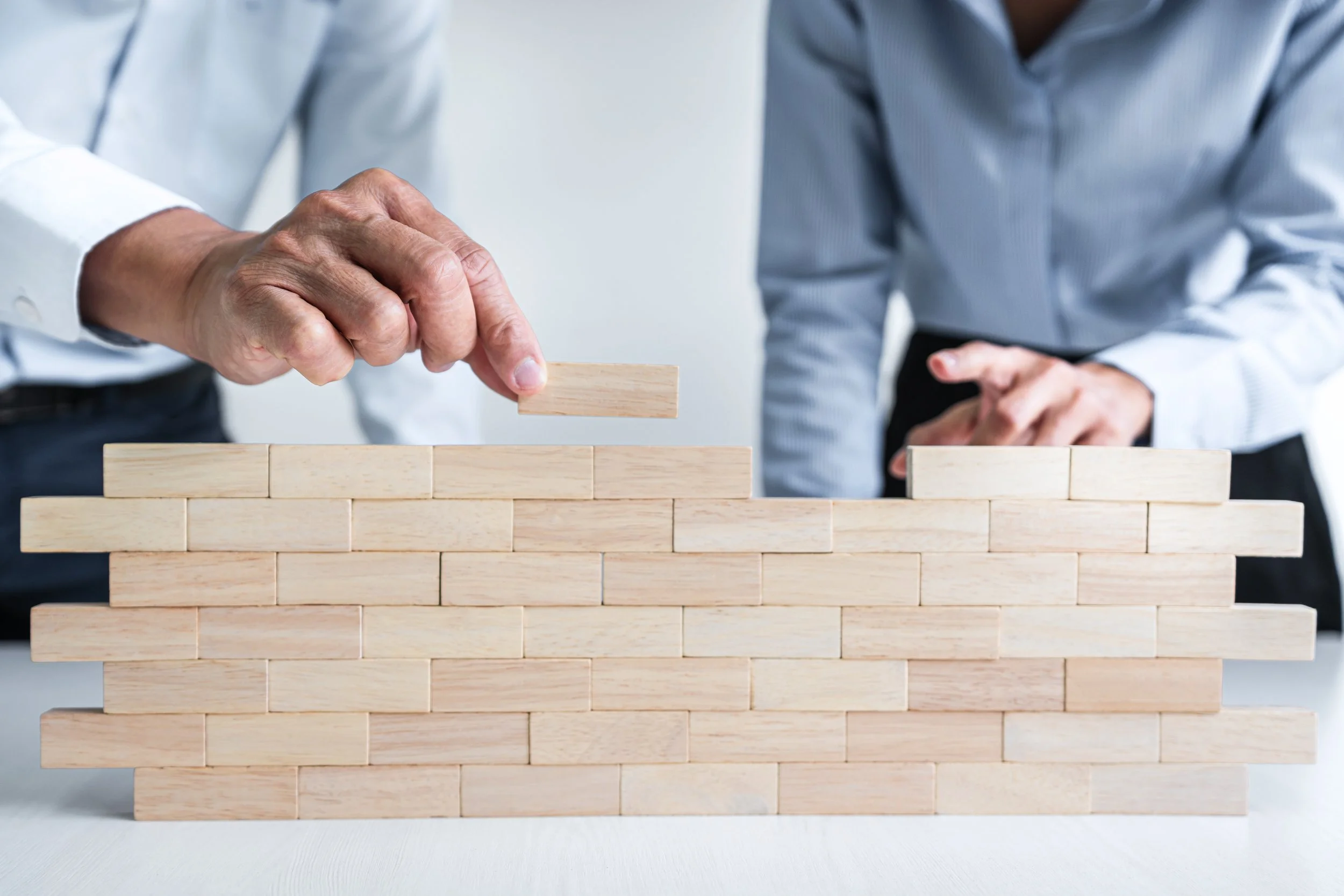 Two people in business attire building a small brick wall with wooden blocks on a white table, one person placing a block on top.