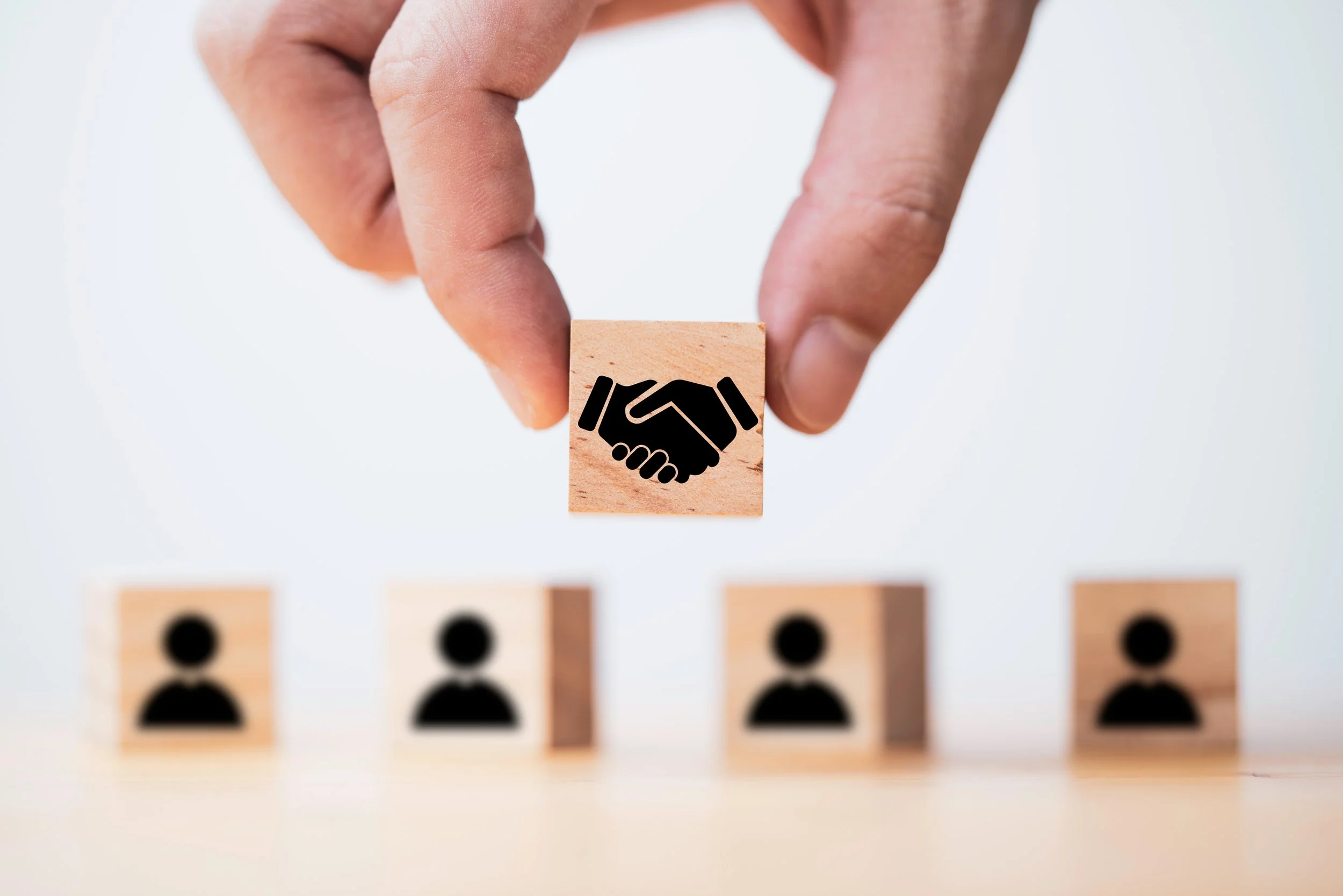 Close-up of a hand holding a wooden block with a handshake icon, with three blurred blocks with person icons on a flat surface in the foreground.