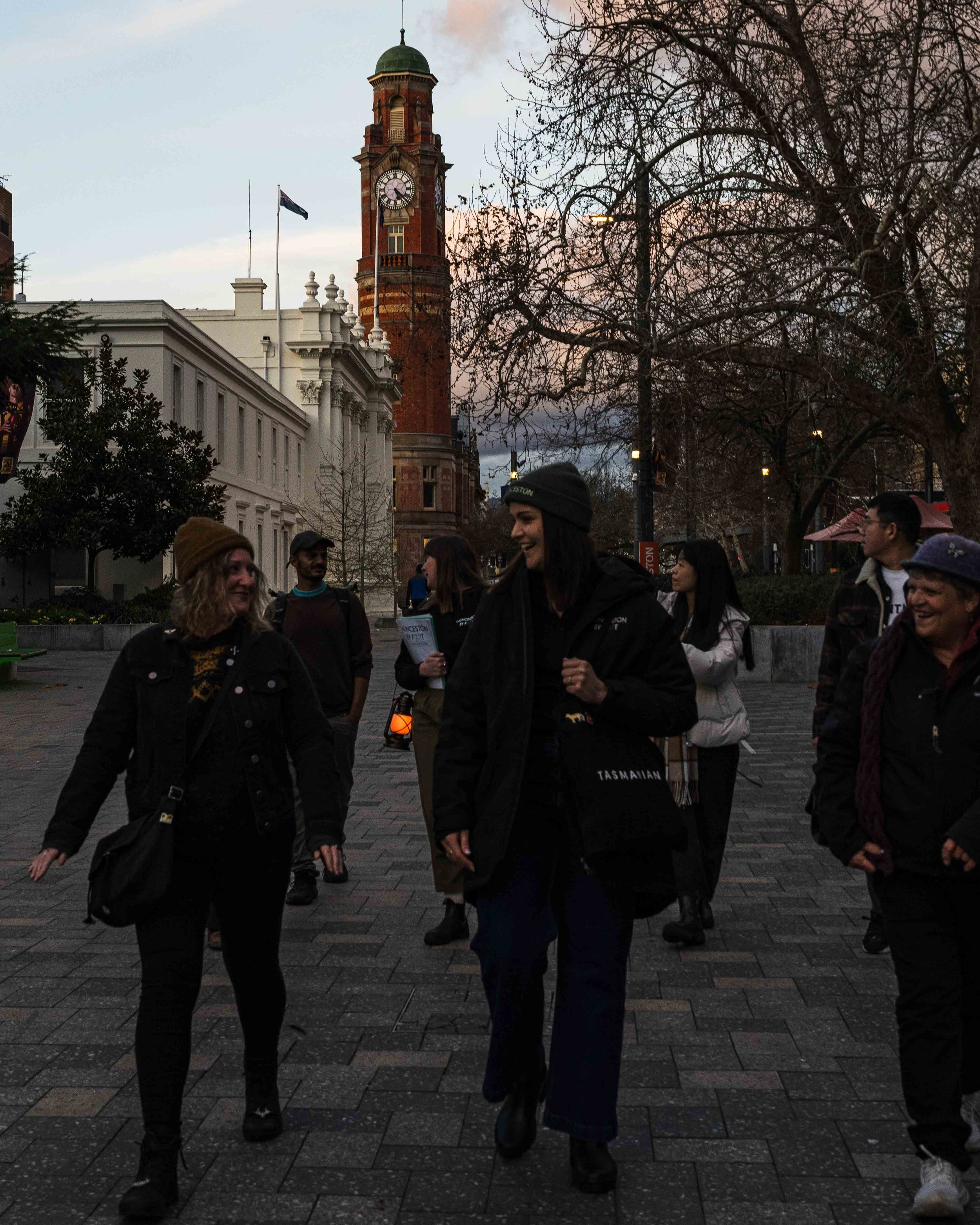 A group of people socializing outdoors during evening with a historic clock tower and leafless trees in the background.
