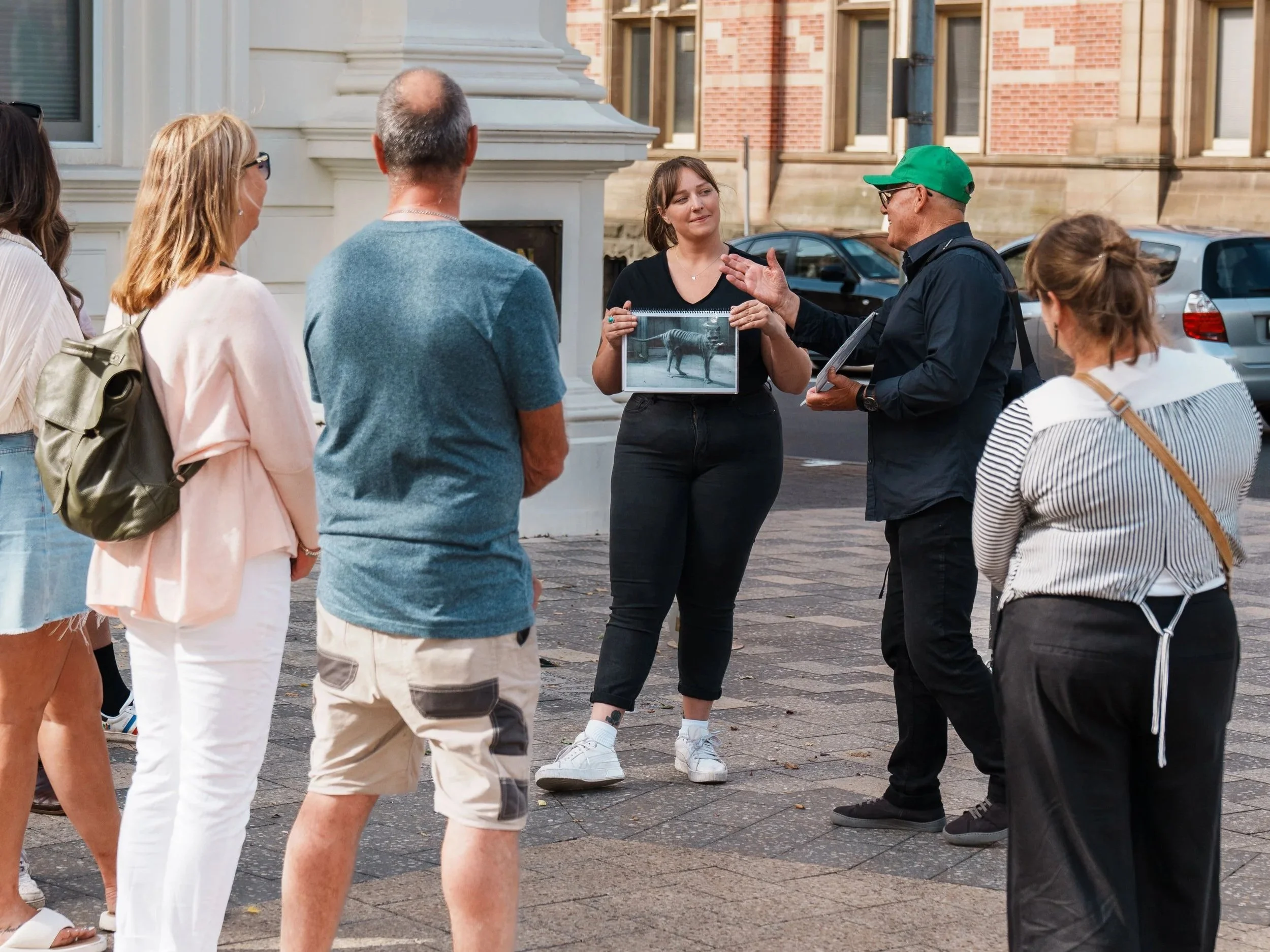 A woman holding a photo of a zebra, speaking to a group of people on a city street with brick buildings and parked cars in the background.