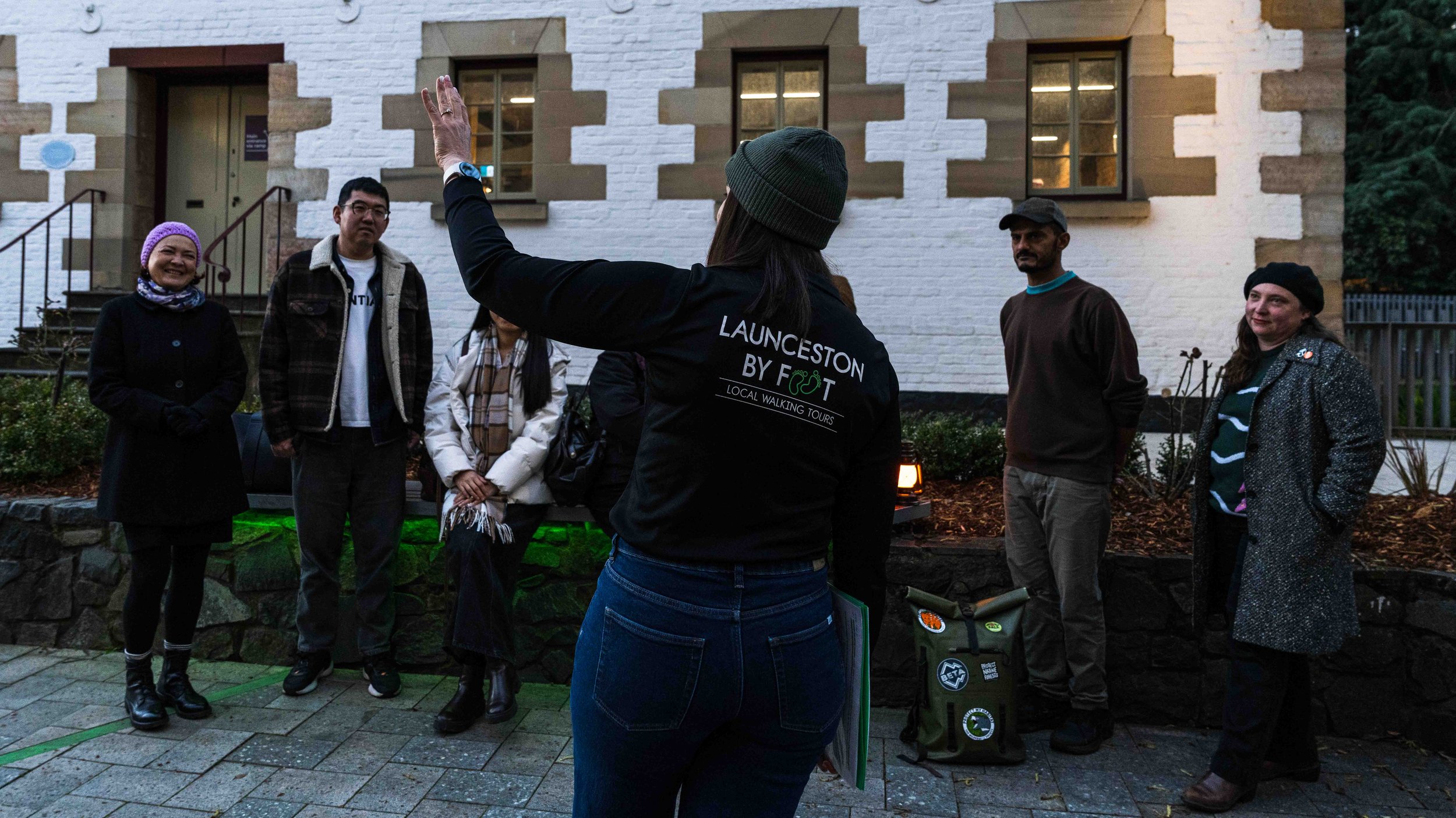 A woman wearing a beanie and a black jacket with the text 'LAUNCESTON BY FOOT' is leading a small group on a walking tour in front of a white brick building with brown trim. The group is listening attentively, dressed in casual outdoor clothing.
