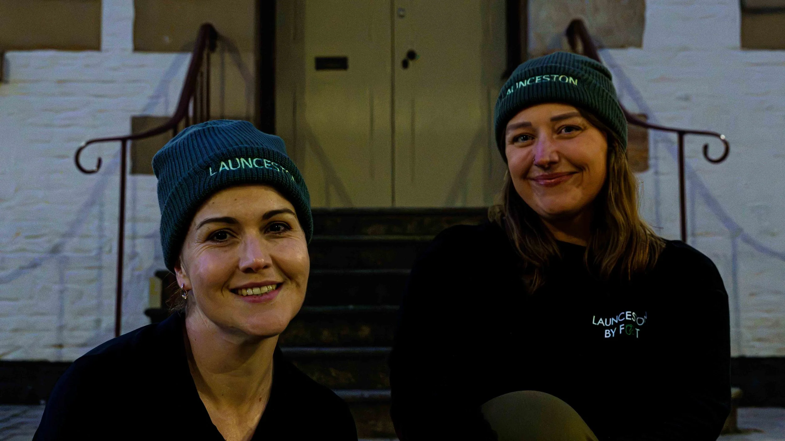 Two women sitting on stairs in front of a building, wearing matching dark beanies with green text, smiling at the camera.