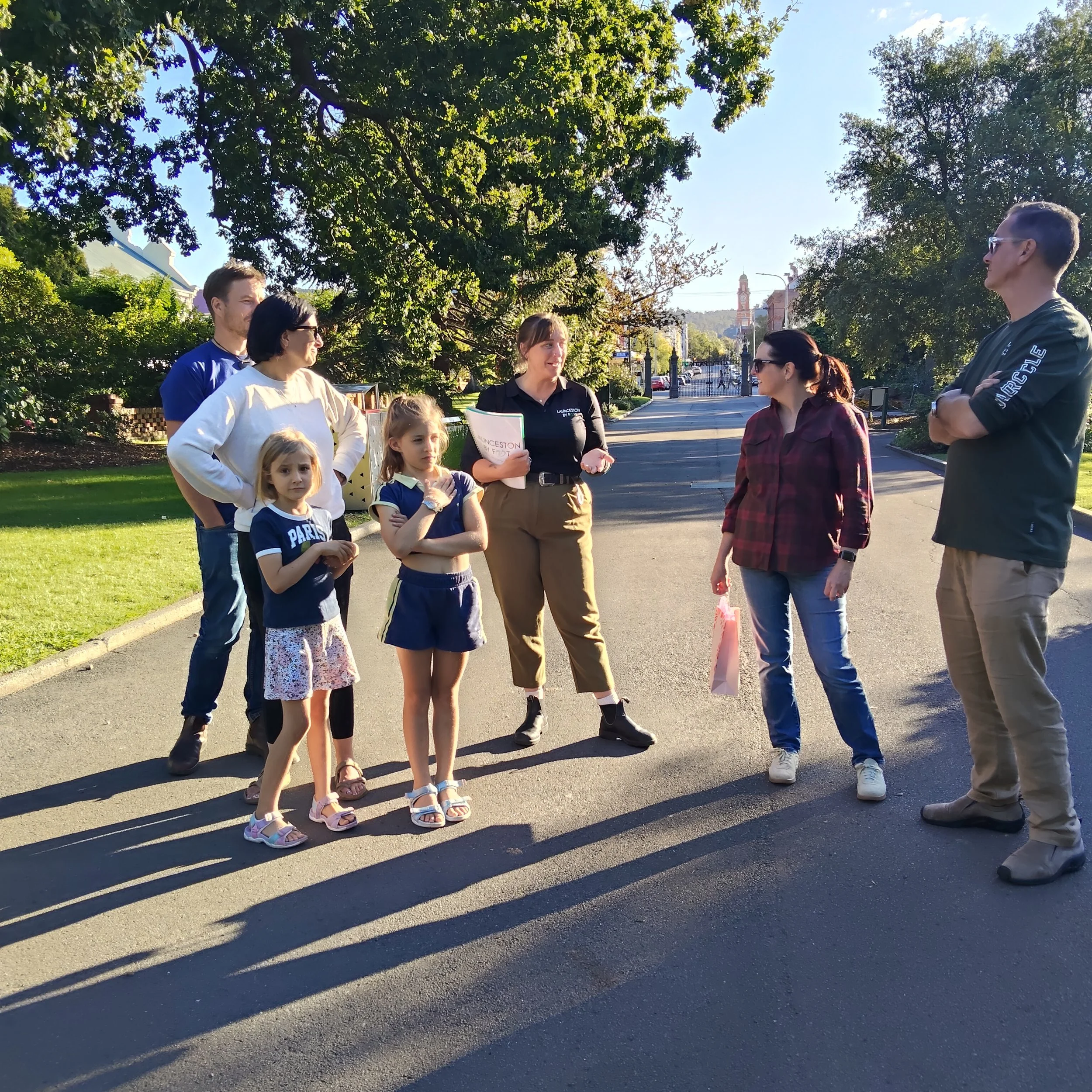 Group of adults and children standing outdoors in a park, engaging in conversation on a sunny day with trees and a building tower in the background.