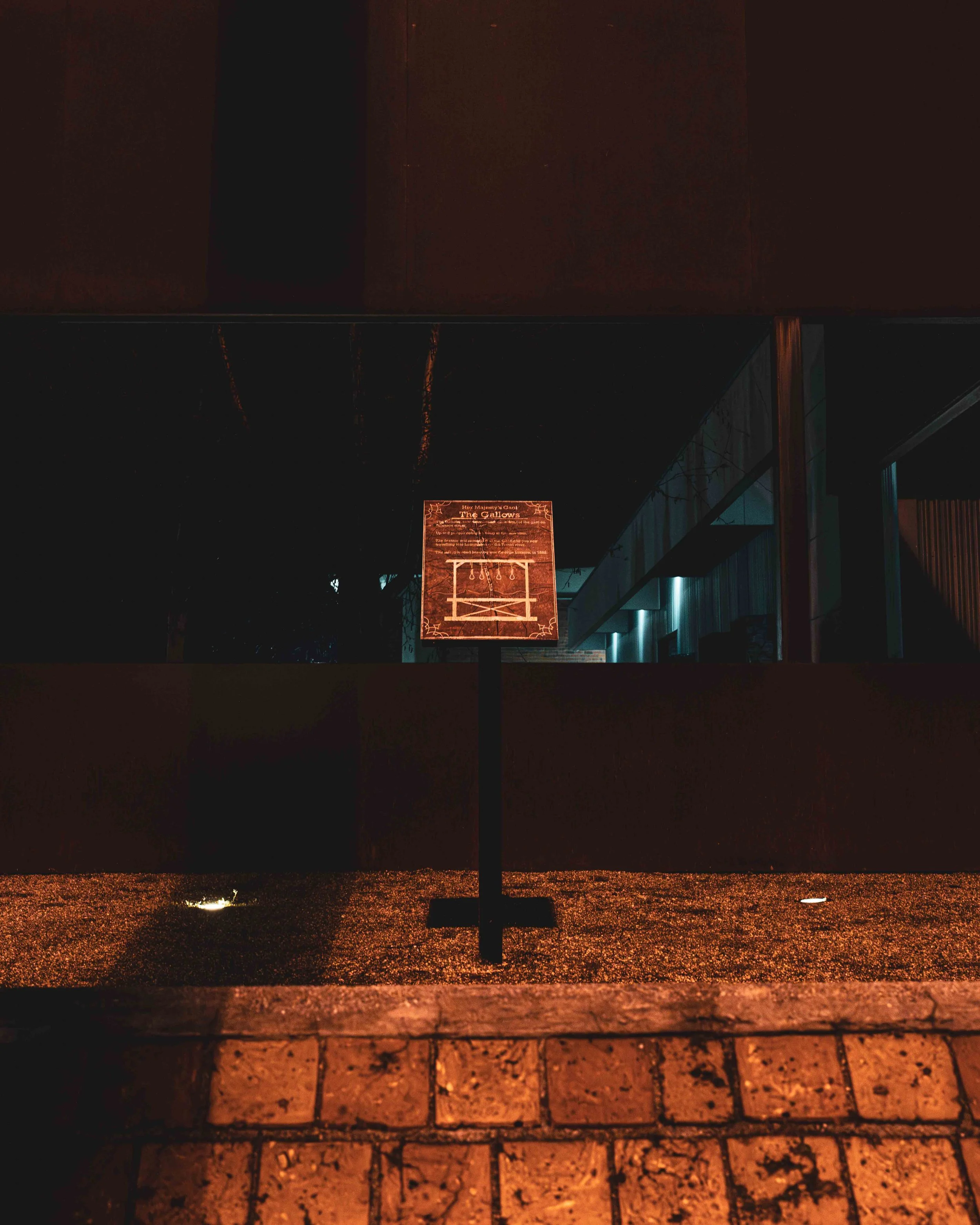 Nighttime scene of a wet street with a metal grate in the foreground and a wall with a sign reading 'The Gallow' illuminated in the distance. The sign features an illustration of a gallows.
